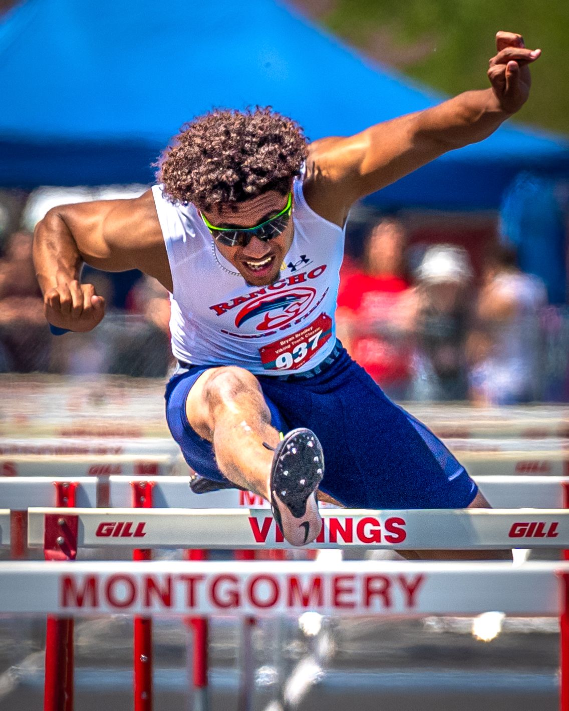 Track athlete clearing hurdles during competition wearing racing uniform and sunglasses.