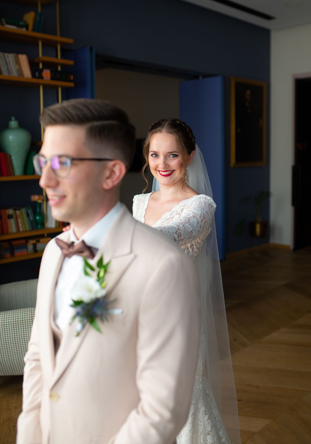 Bride in lace wedding dress and veil smiles at groom during indoor first look moment.