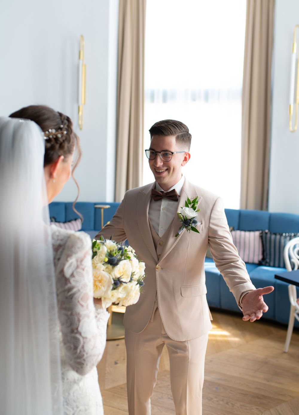 Groom in beige suit reacts with joy seeing bride in lace dress during first look.