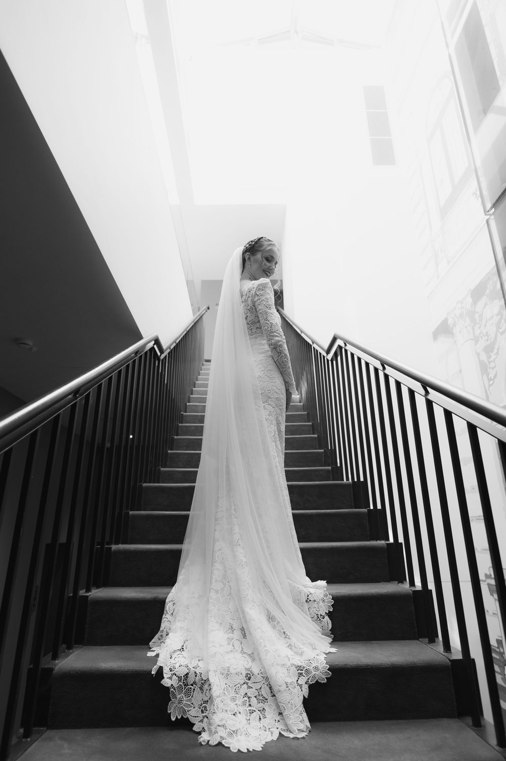 Black and white photo of bride in lace gown and long veil ascending grand staircase.