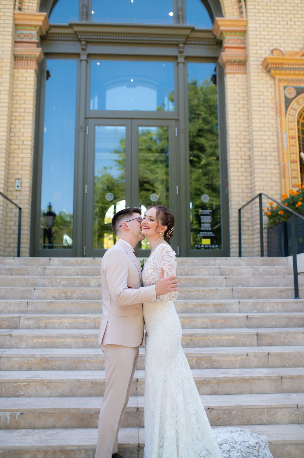 Bride and groom embrace and kiss on steps of ornate building exterior on wedding day.