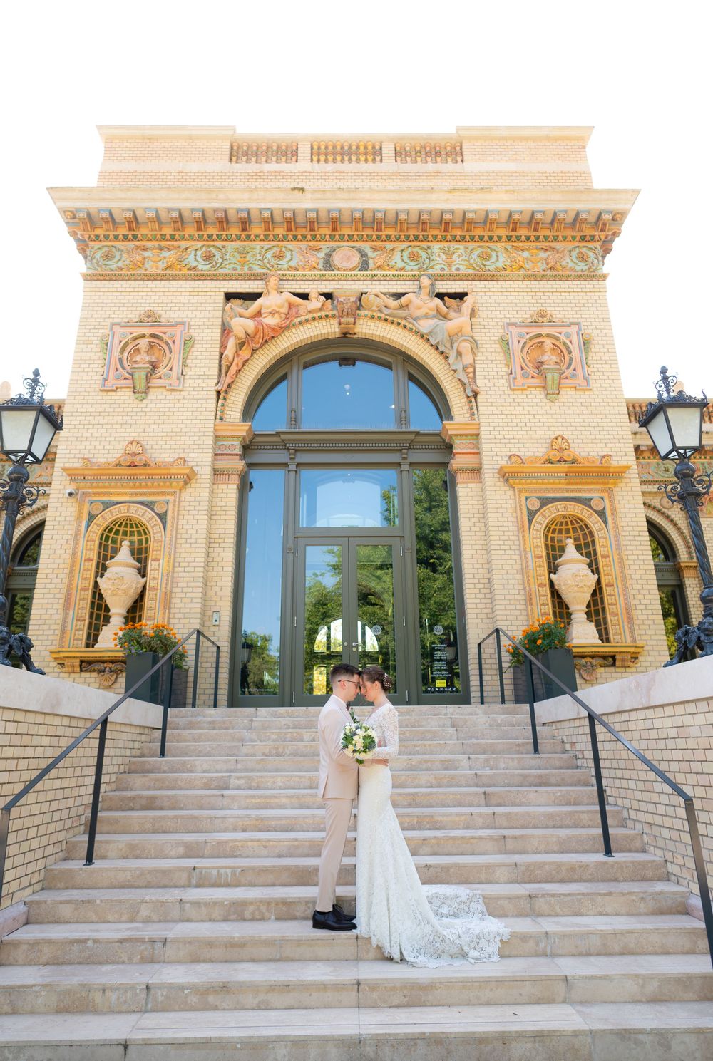 Bride and groom kiss on steps of ornate historic building with decorative facade.