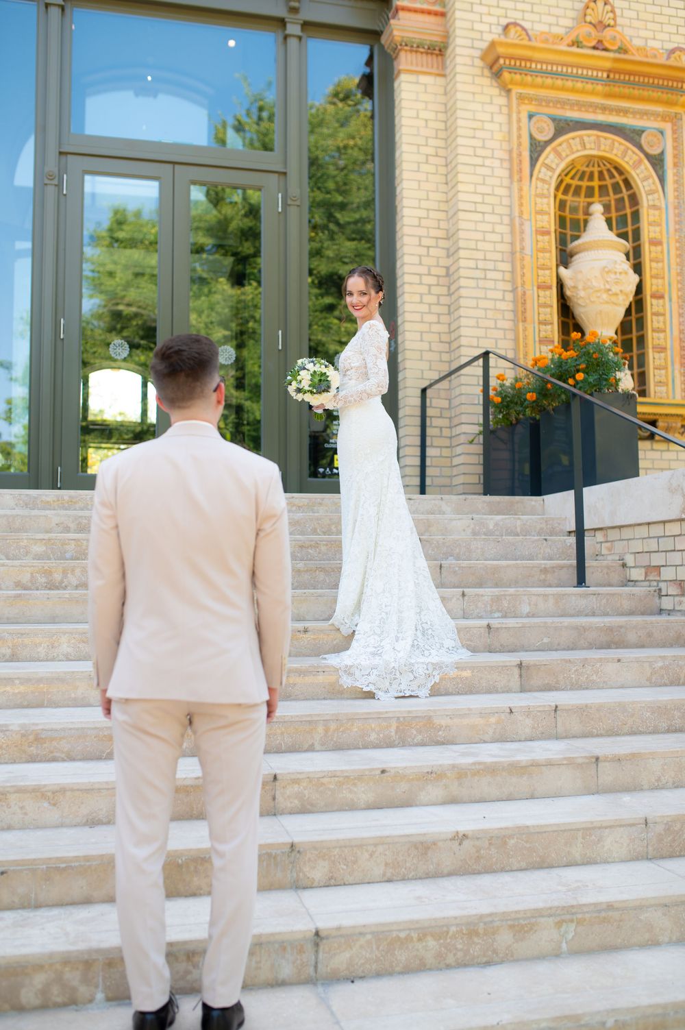 Bride in lace gown looks back at groom standing on steps of elegant building exterior.