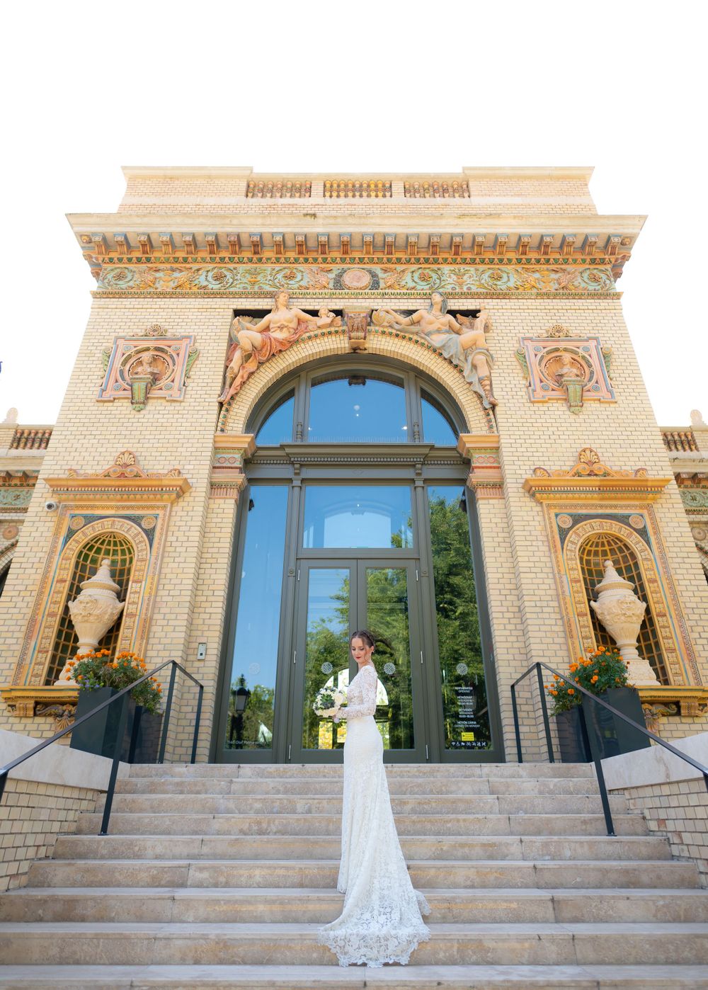 Bride in fitted lace wedding gown poses alone on steps of ornate historic building.
