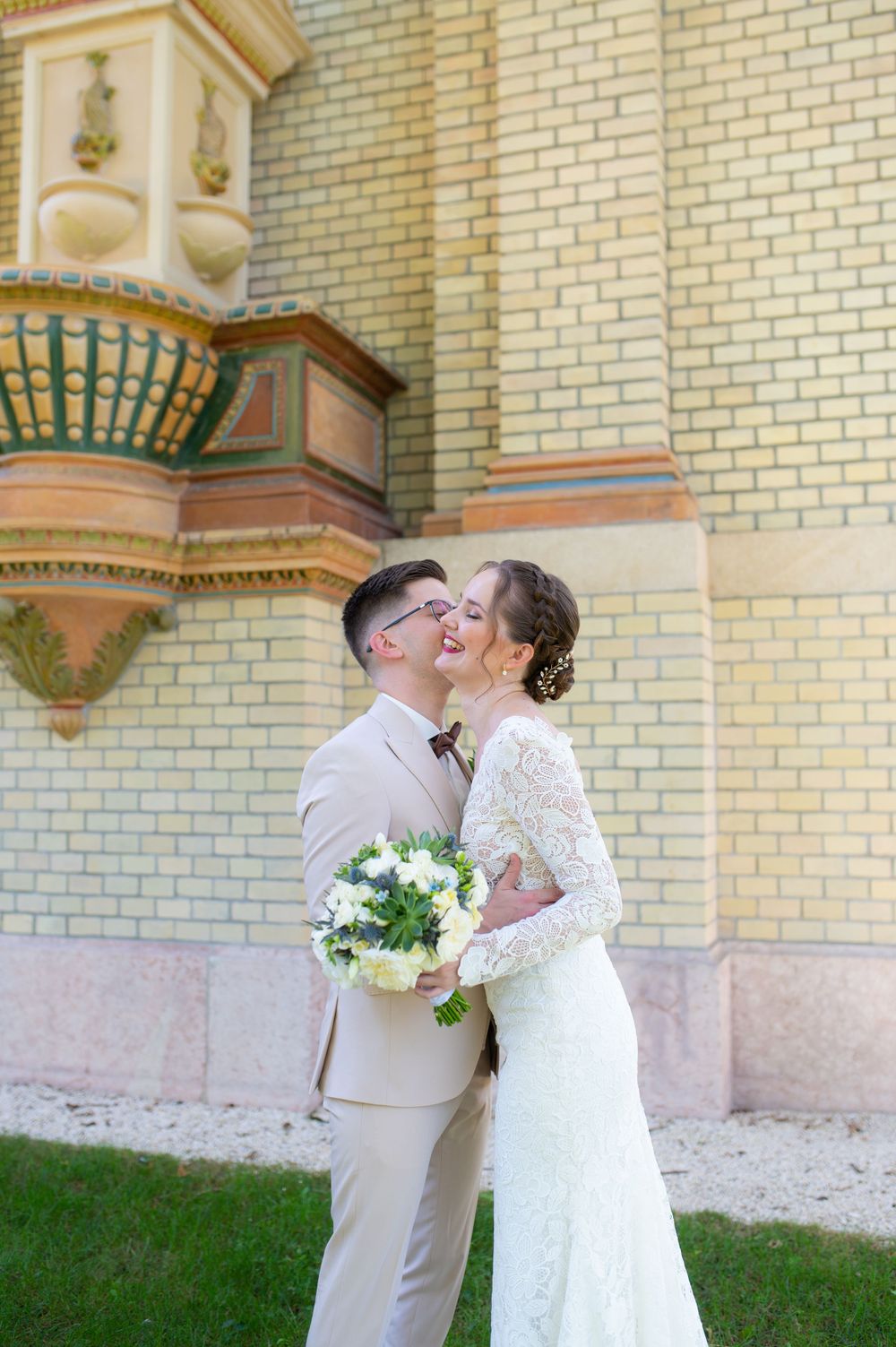 Bride and groom sharing a romantic kiss beside an ornate brick building, bride holding white floral bouquet.