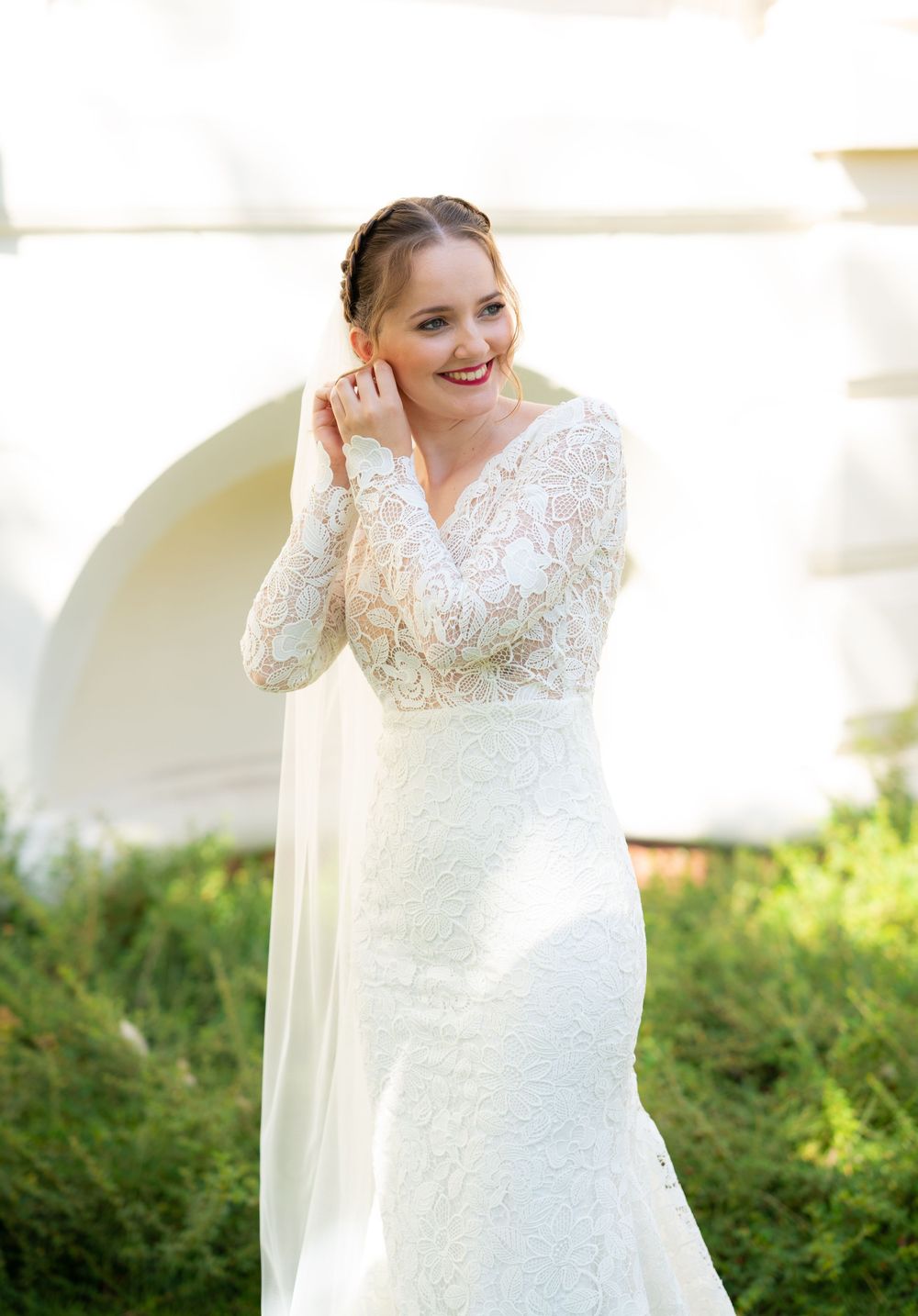 Bride in long-sleeve lace wedding gown with flowing veil smiling outdoors in bright sunlight.