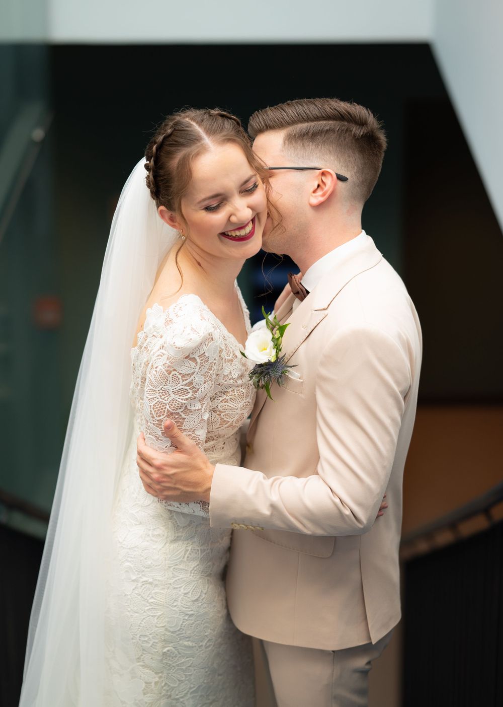 Groom in beige suit whispering to laughing bride in lace wedding dress during portrait session.