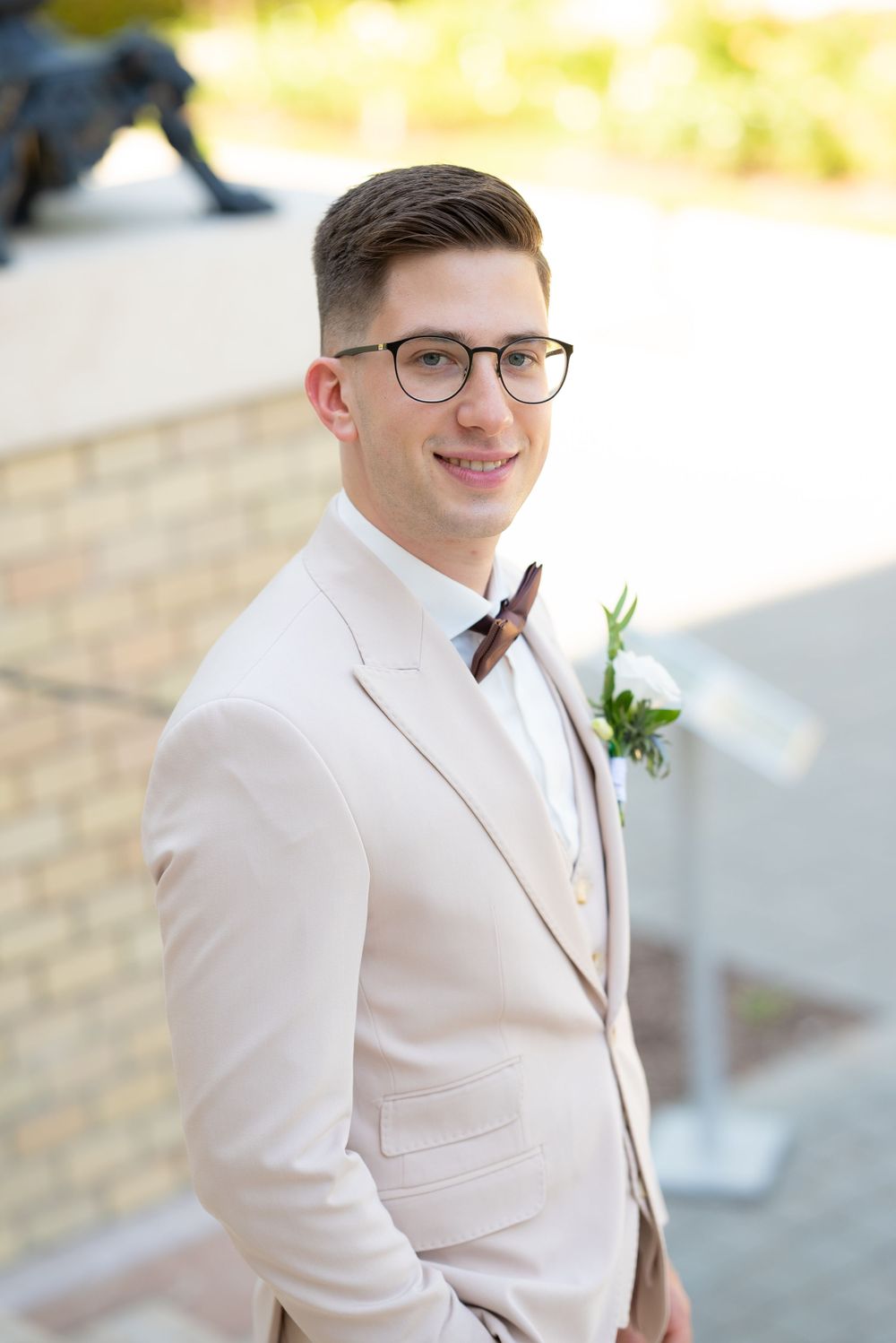 Handsome groom in beige suit with wood bow tie and boutonniere smiling outdoors on wedding day.