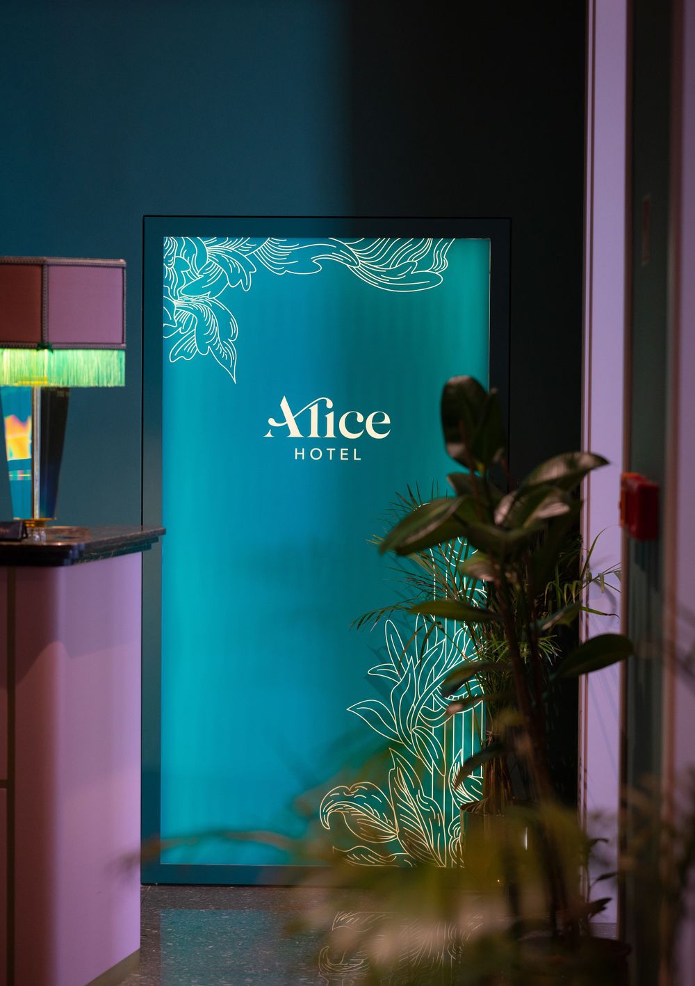 Glowing teal Alice Hotel sign with floral design in a stylishly decorated hotel lobby.