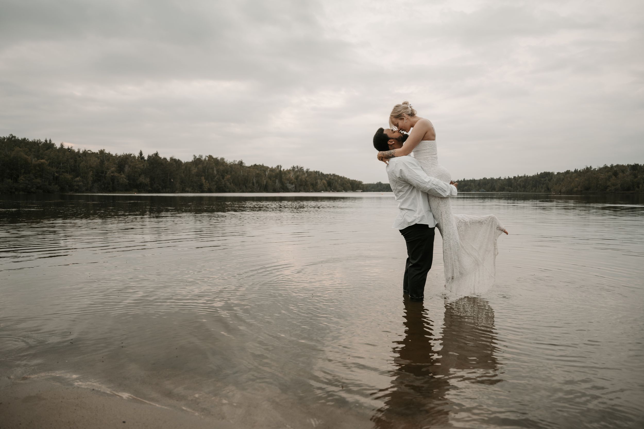A couple shares a romantic embrace while standing in shallow lake water during a cloudy day, the bride's dress trailing in the water.