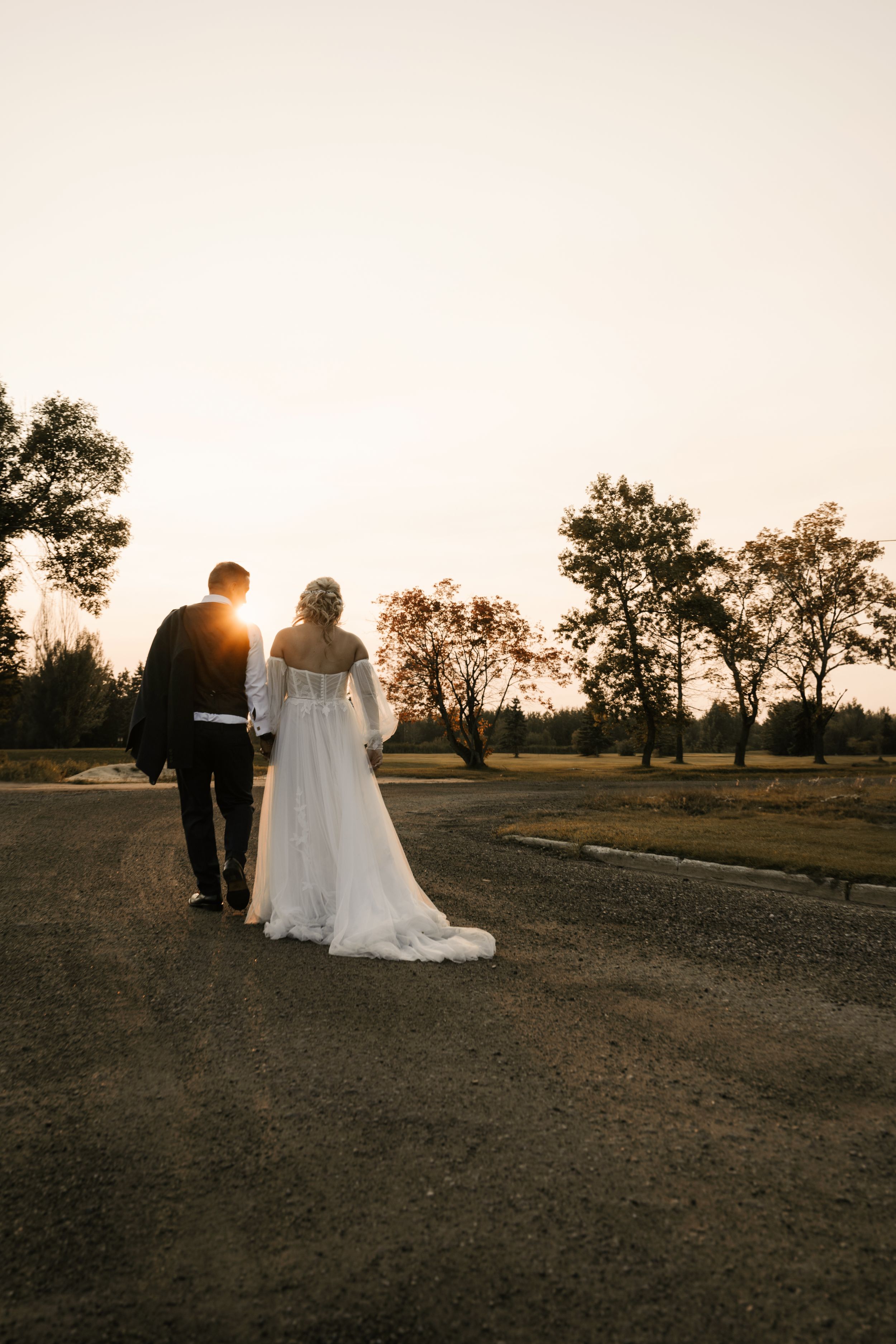 A couple in wedding attire walks along a dirt path at sunset with silhouetted trees in the background.