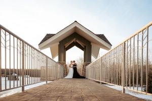A wedding couple stand in silhouette beneath a covered bridge walkway with metal railings against a bright sky.