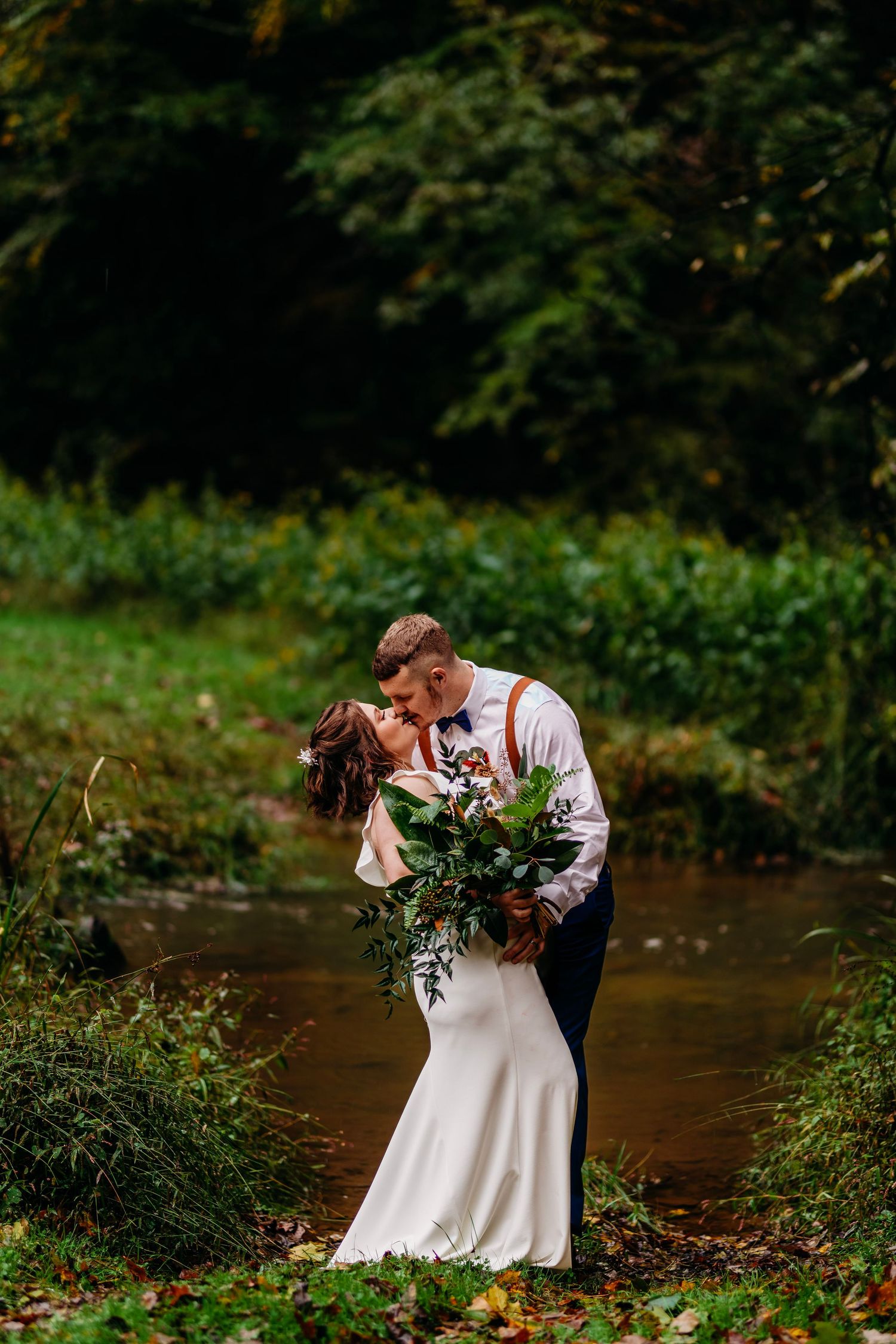 Bride and groom share a romantic kiss by a creek surrounded by lush greenery.