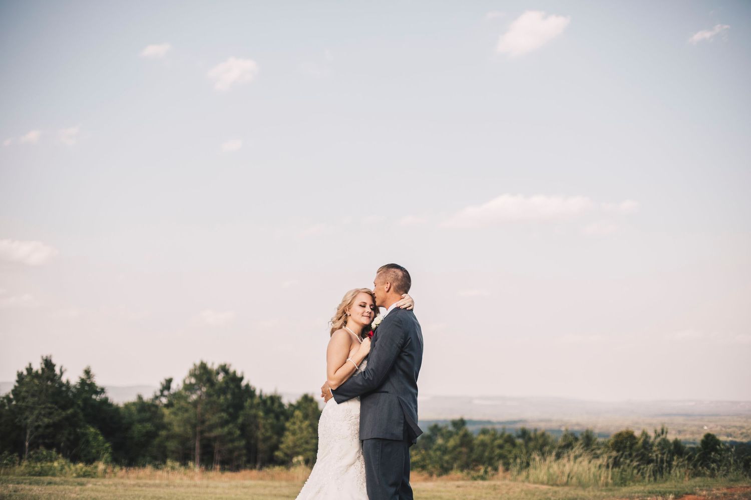 Bride and groom embracing in open field with trees and blue sky in background.