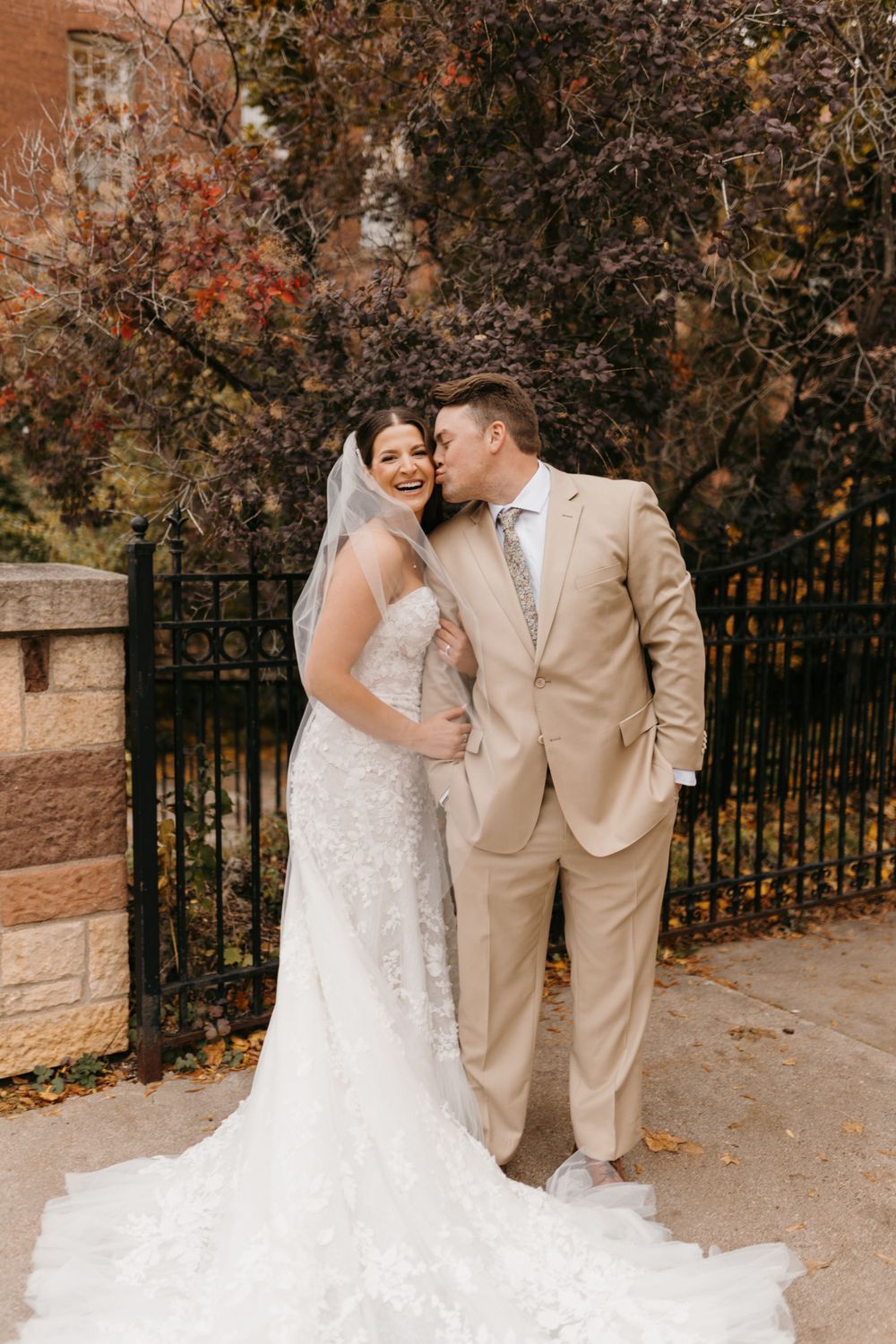 Bride in lace gown and groom in tan suit share a joyful moment by iron fence with autumn foliage.