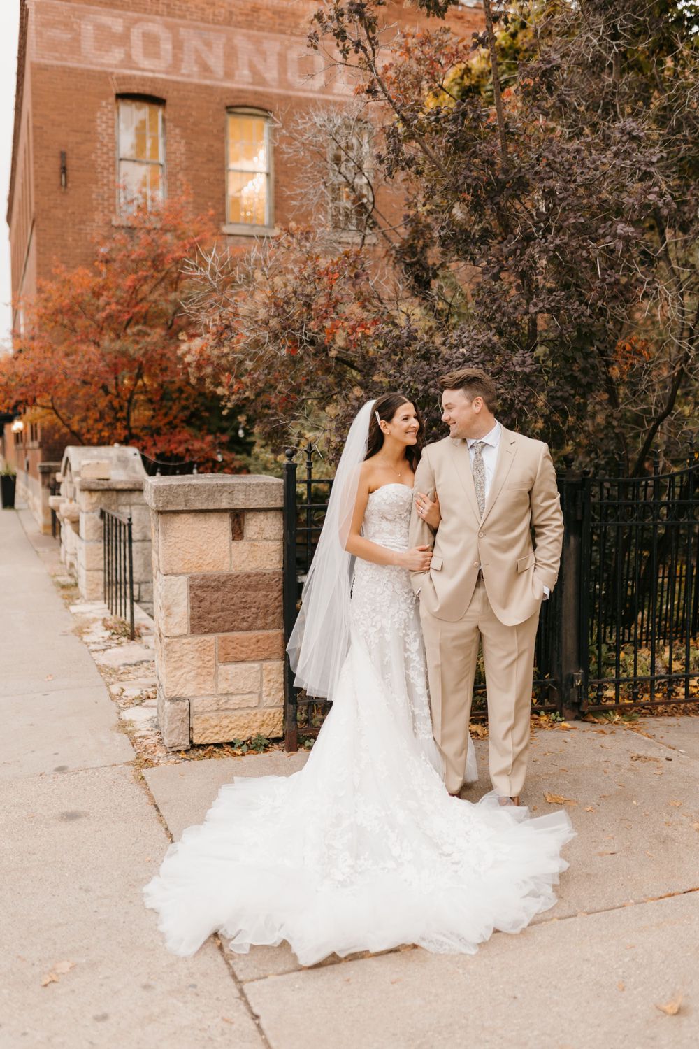Bride and groom share a loving glance on sidewalk with autumn foliage and brick building backdrop.