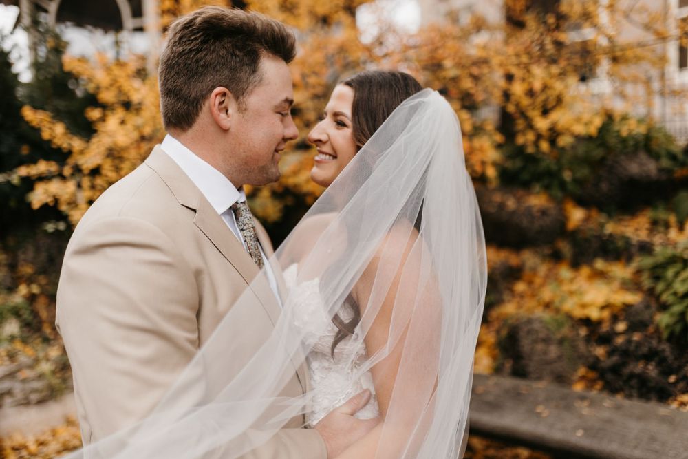 Bride and groom share intimate moment wrapped in flowing veil with golden autumn trees behind them.