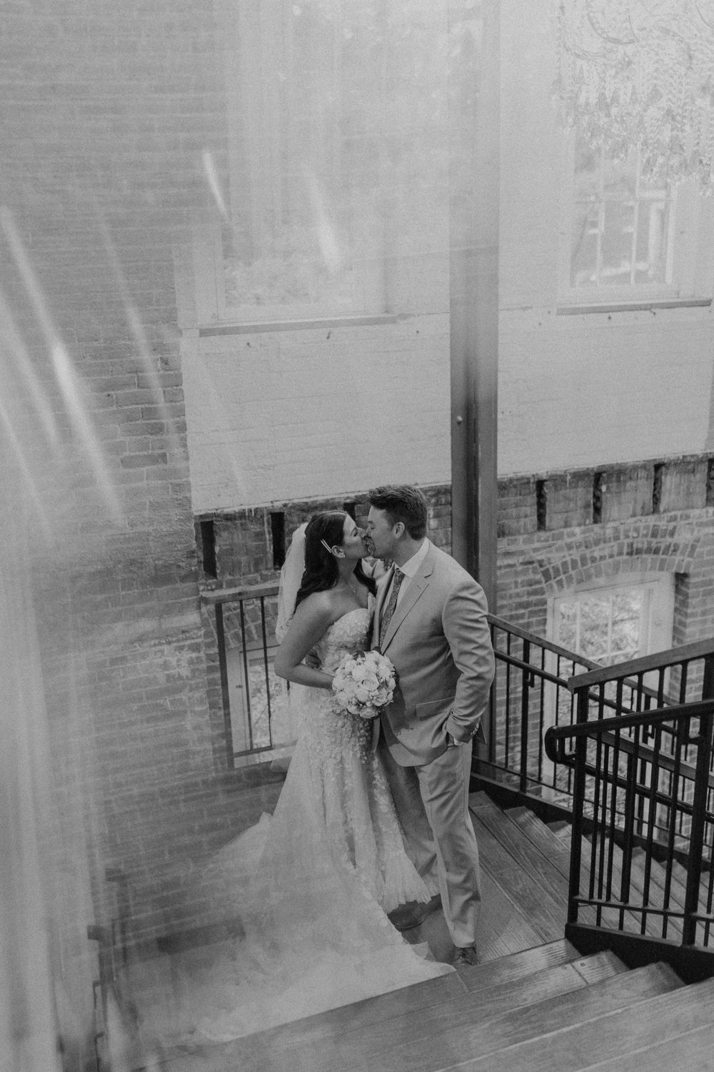 Black and white photo of bride and groom kissing on staircase inside rustic brick wedding venue.