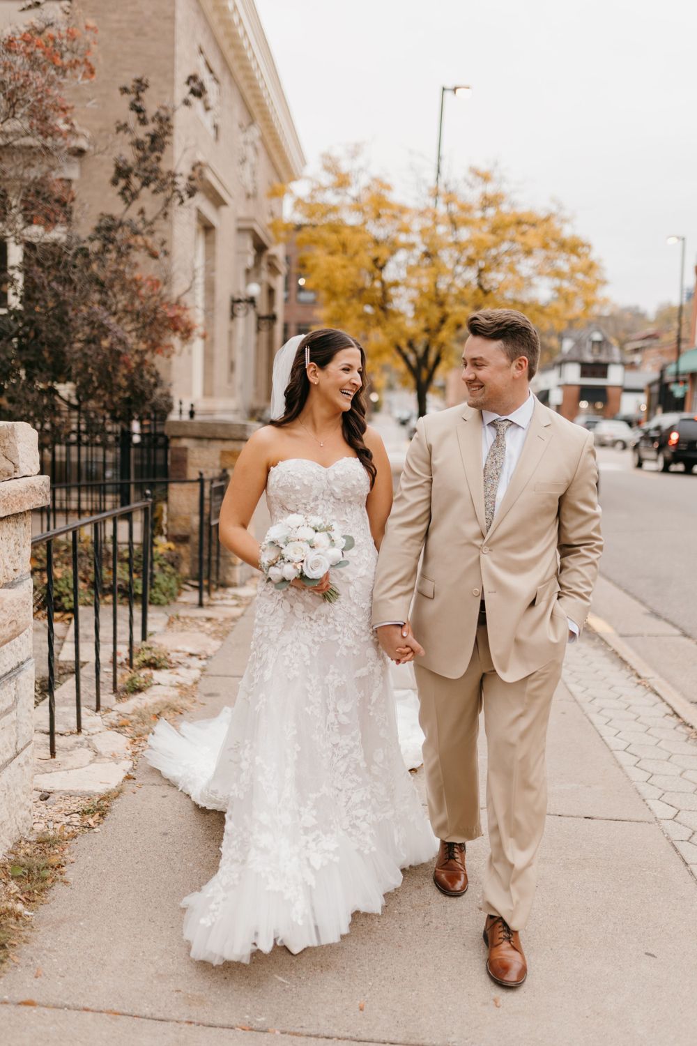 Bride holding white bouquet and groom in tan suit walk hand-in-hand on urban sidewalk in autumn.