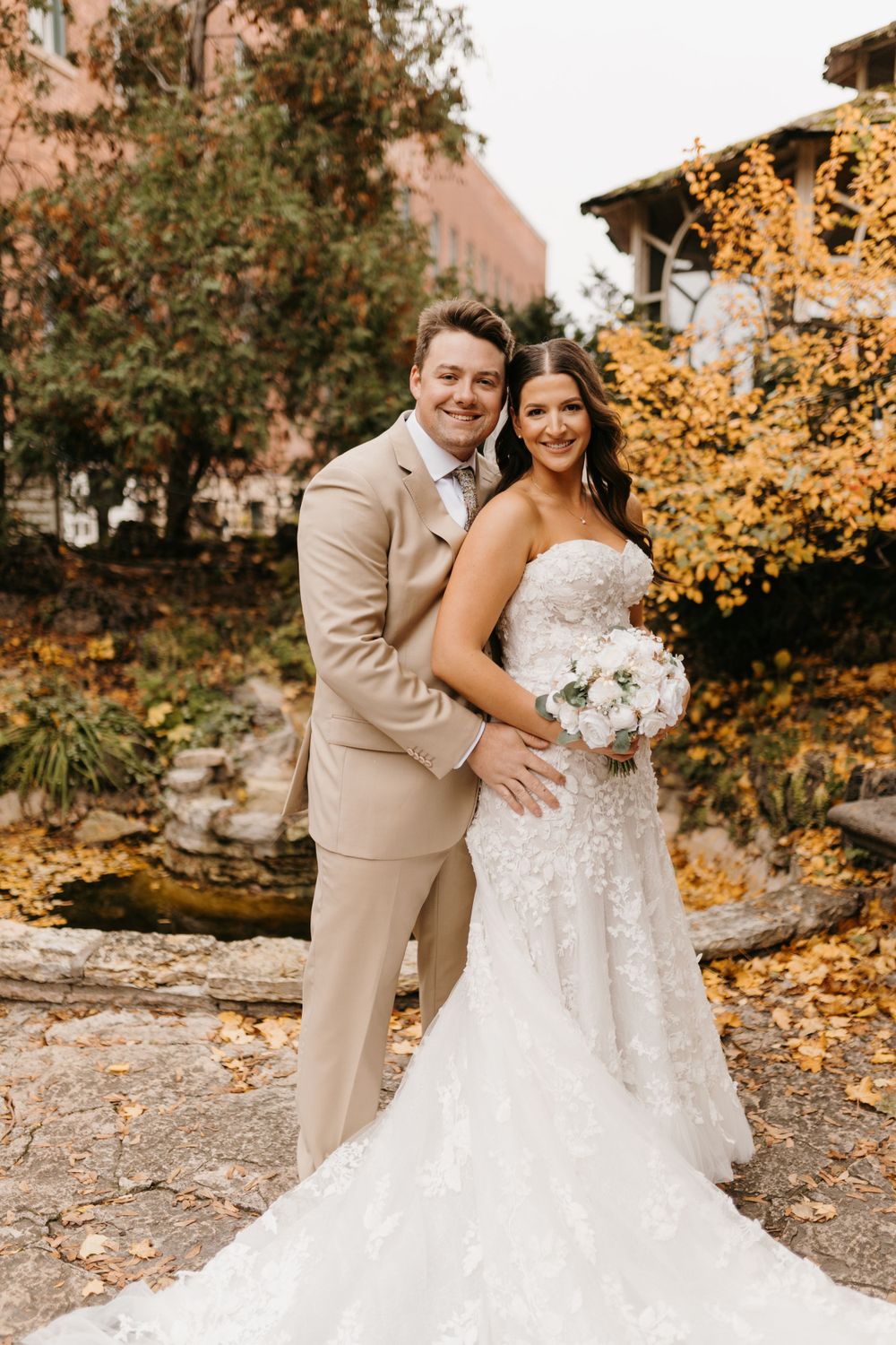 Groom in tan suit embraces bride holding white bouquet surrounded by golden autumn leaves.