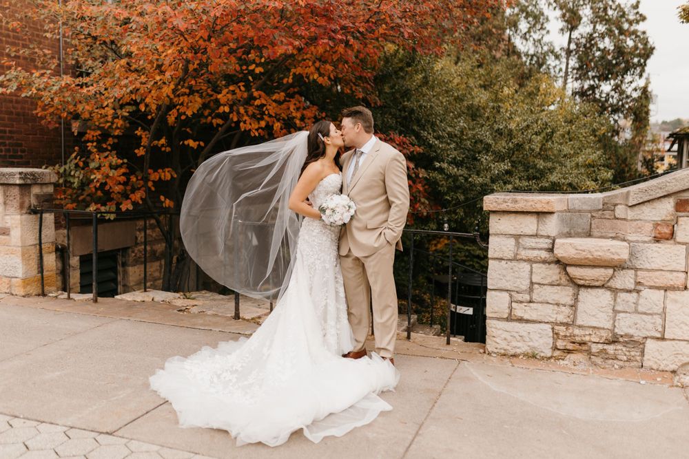 Bride and groom share a kiss outdoors with stunning red fall foliage backdrop, veil flowing in wind.