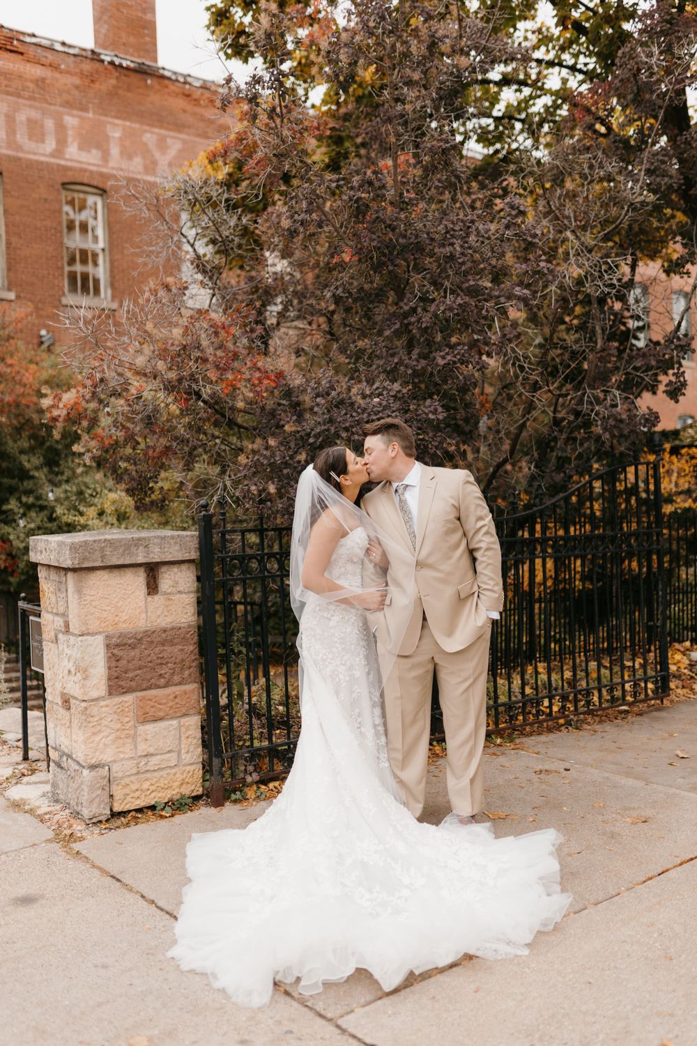 Wedding couple shares a kiss near iron fence with colorful fall trees and brick building backdrop.