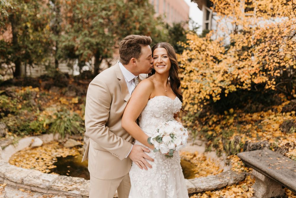 Groom whispers to smiling bride holding white bouquet with golden fall foliage in background.