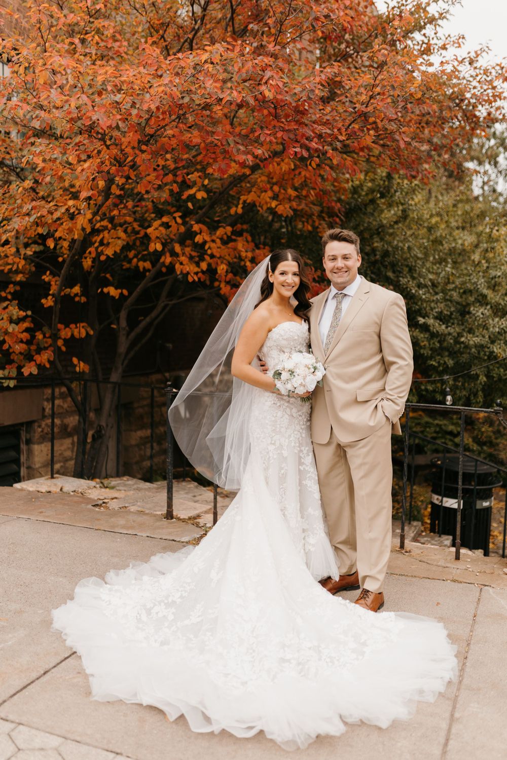Bride in lace gown with veil and groom in tan suit pose before vibrant red autumn foliage.