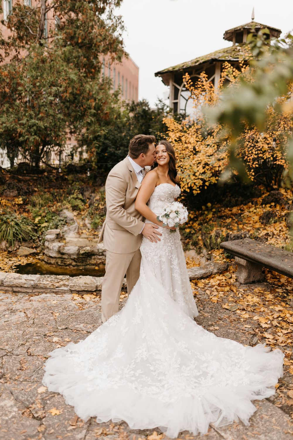 Groom in tan suit embraces laughing bride in lace gown amid golden autumn garden setting.
