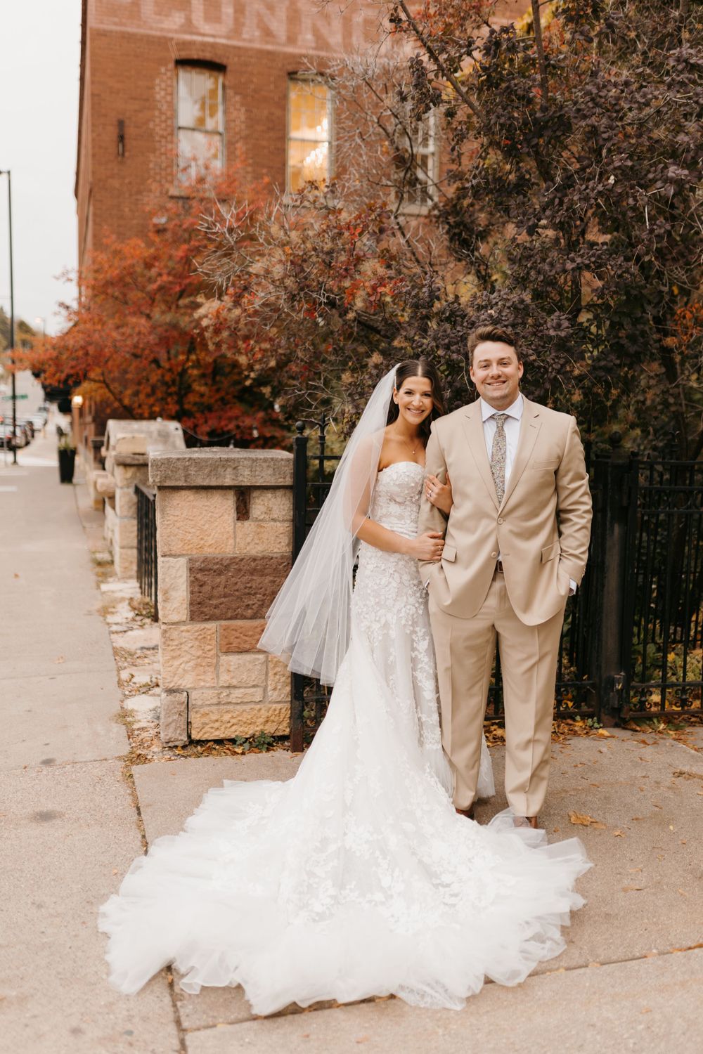 Bride in lace gown with veil and groom in tan suit pose on sidewalk by brick building.