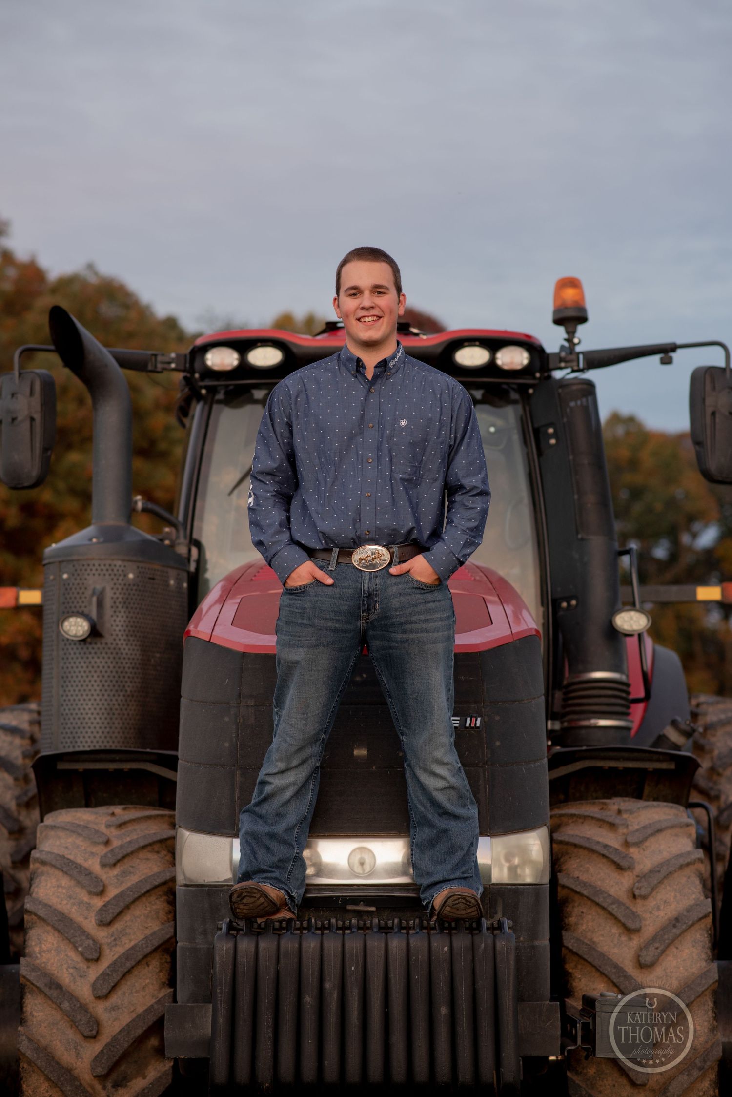 Person stands proudly on the hood of red farm tractor with autumn scenery behind.