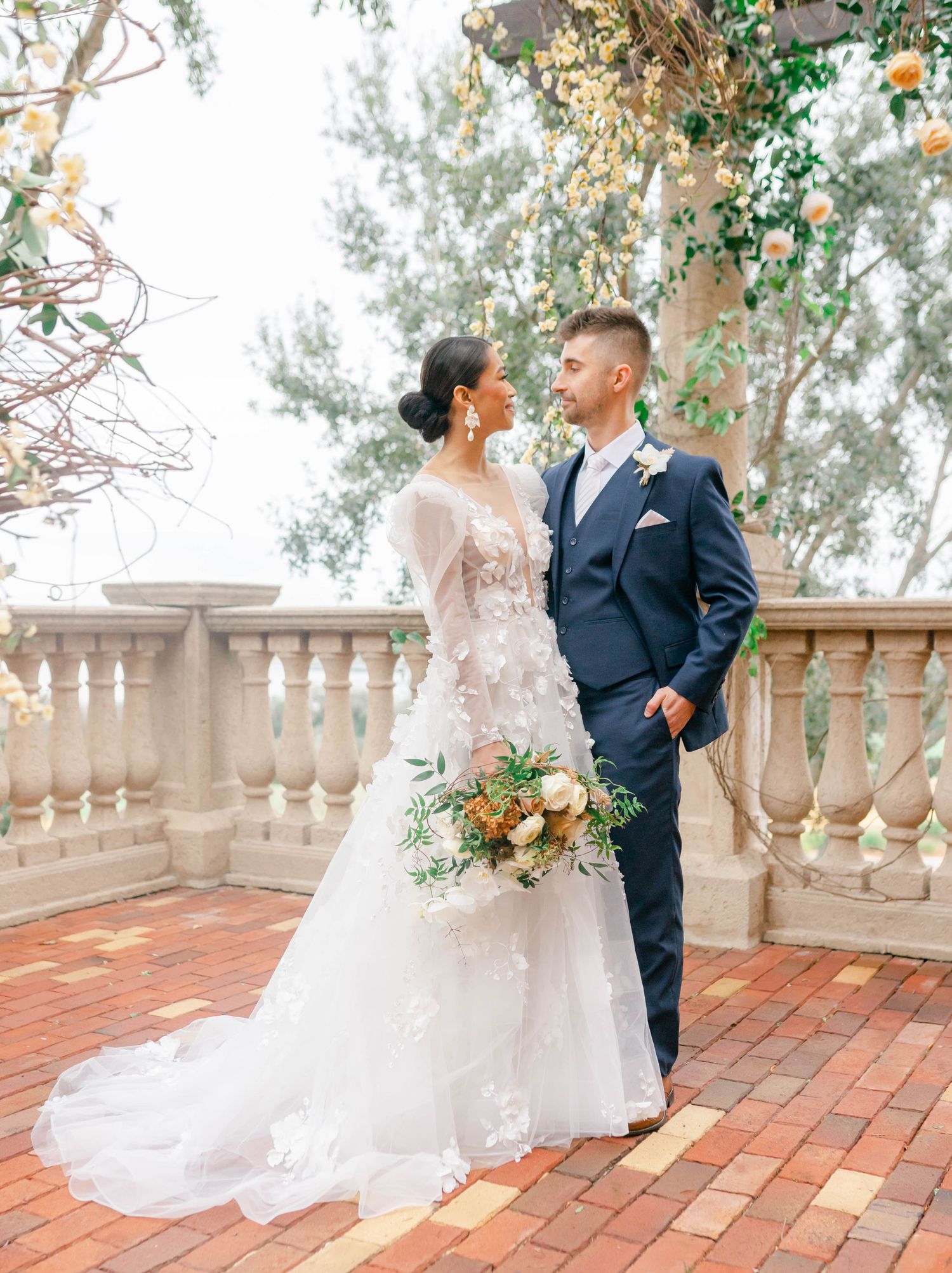 A bride and groom stand together on a brick patio, surrounded by lush greenery.