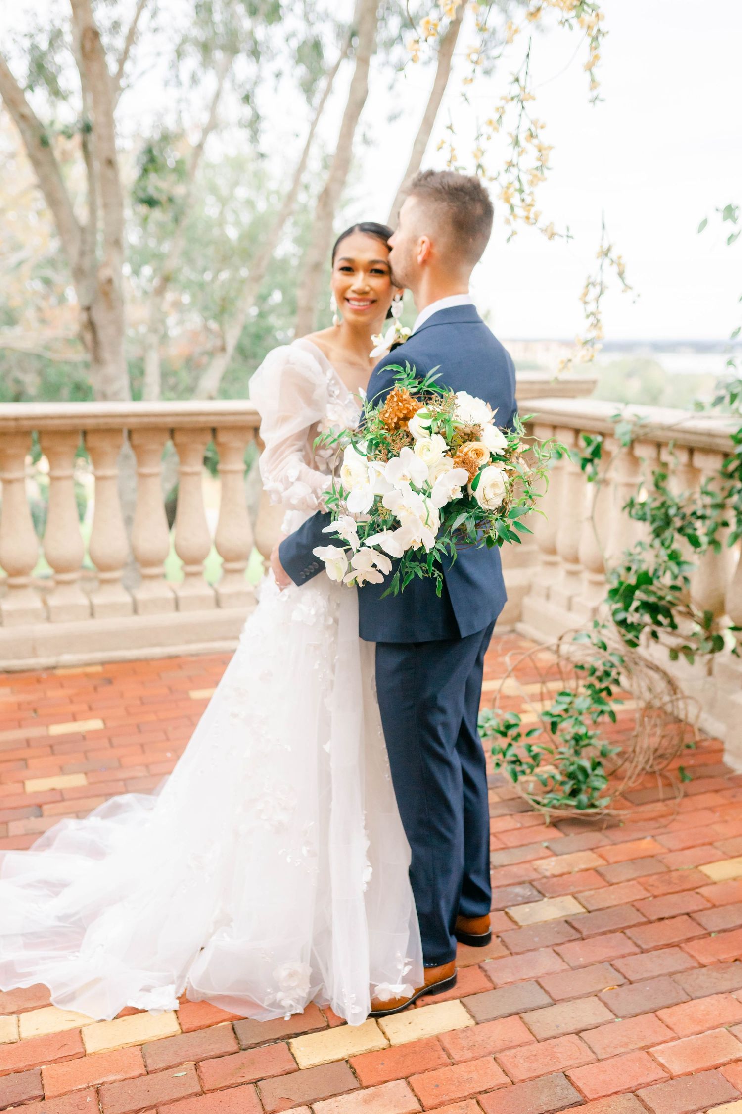 A bride and groom stand together on a brick patio, surrounded by flowers and greenery.