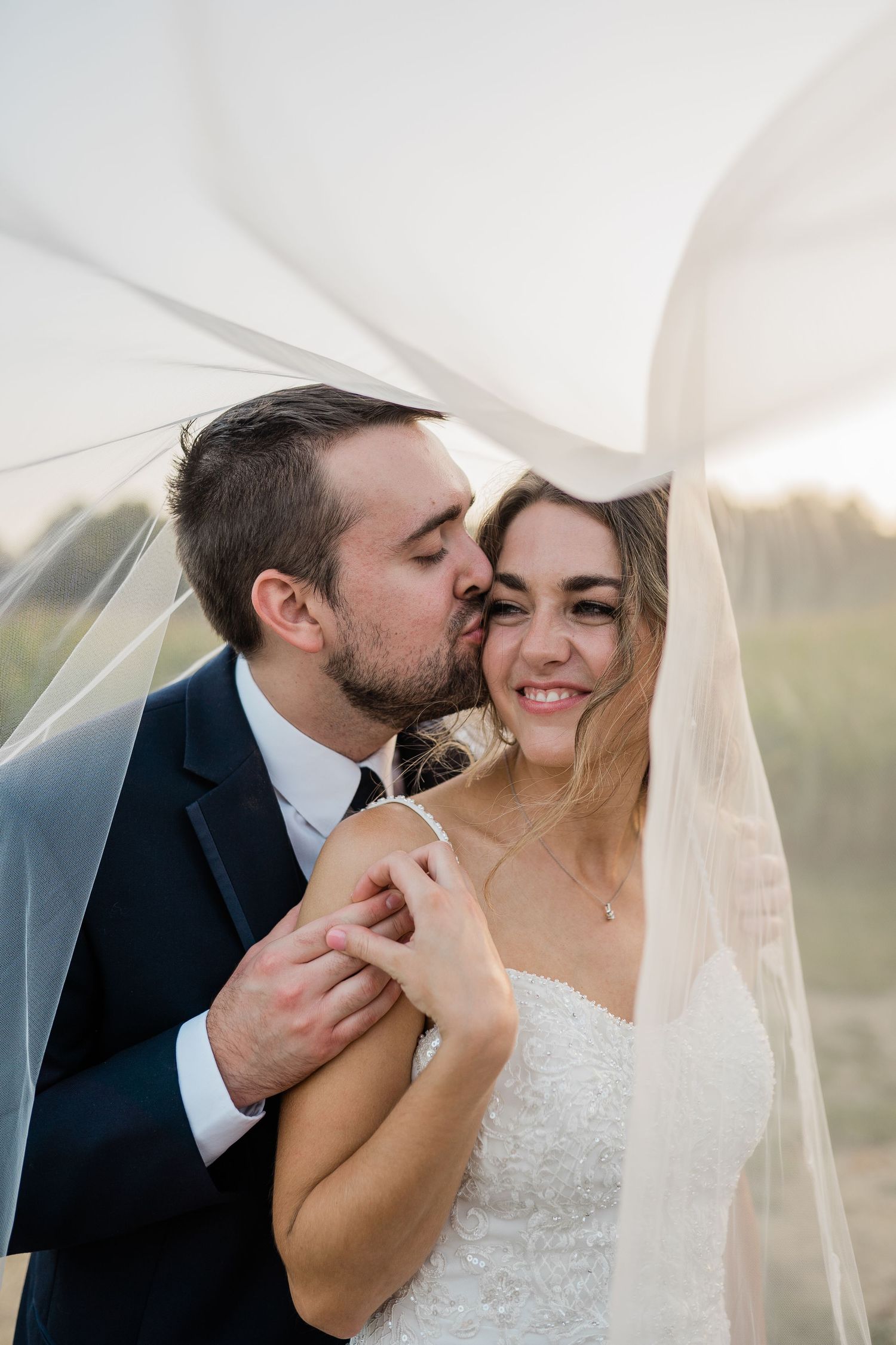 A romantic wedding moment captured under flowing white veil at sunset.