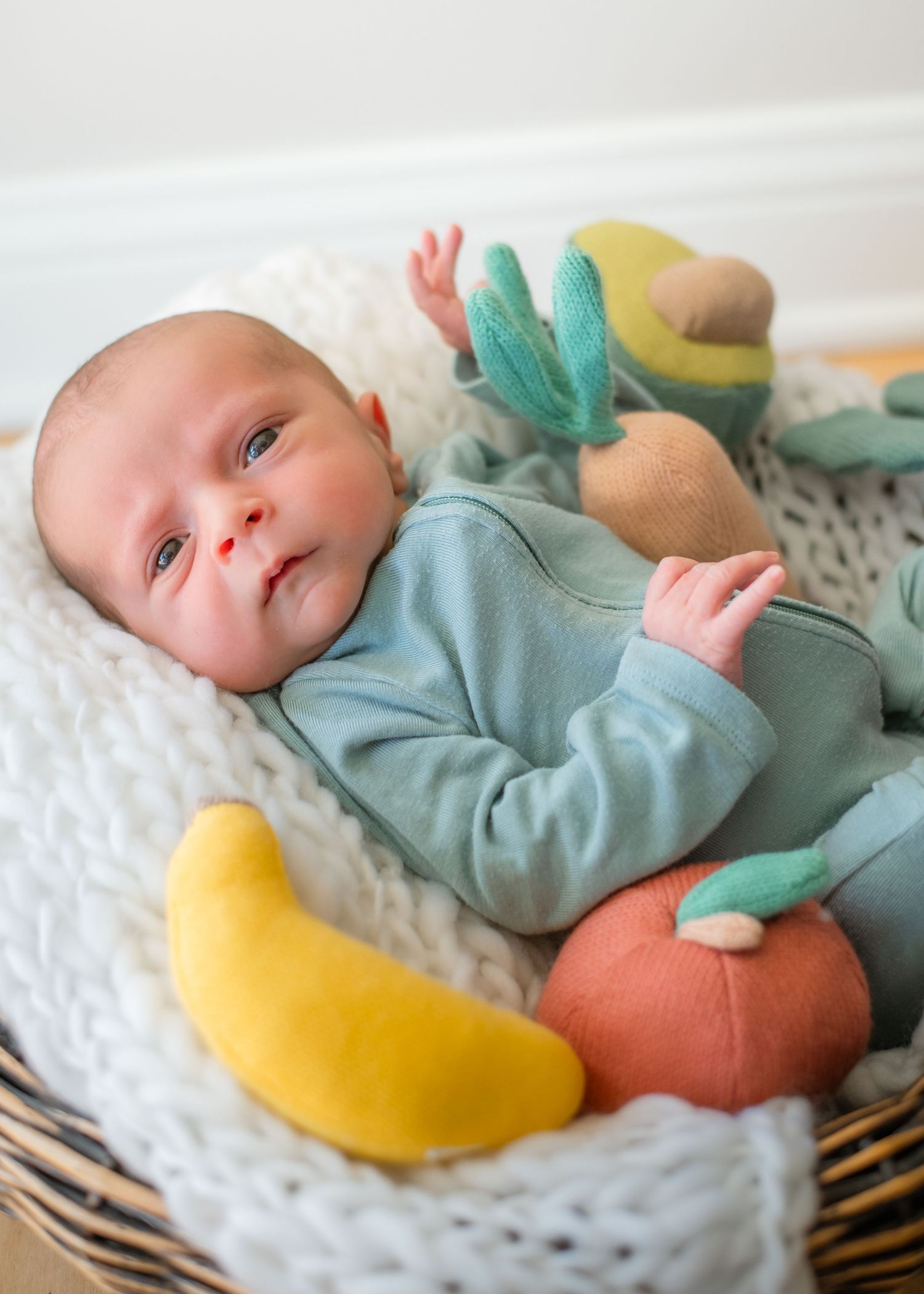 Baby in mint green outfit lying on white blanket with colorful plush toys.