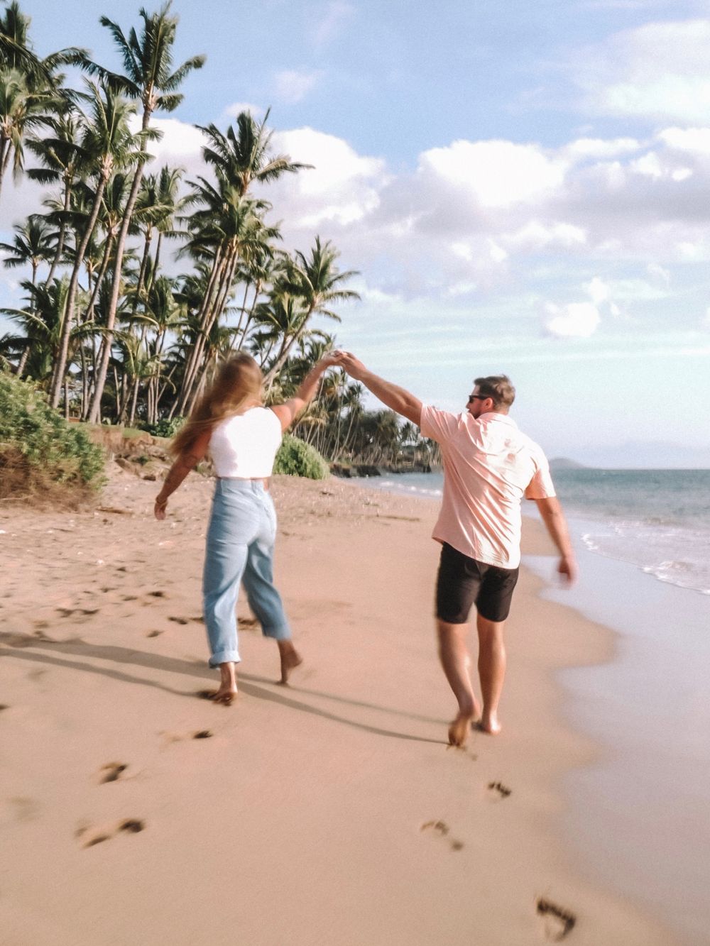 Two people walking and dancing on a palm-lined beach with ocean views.