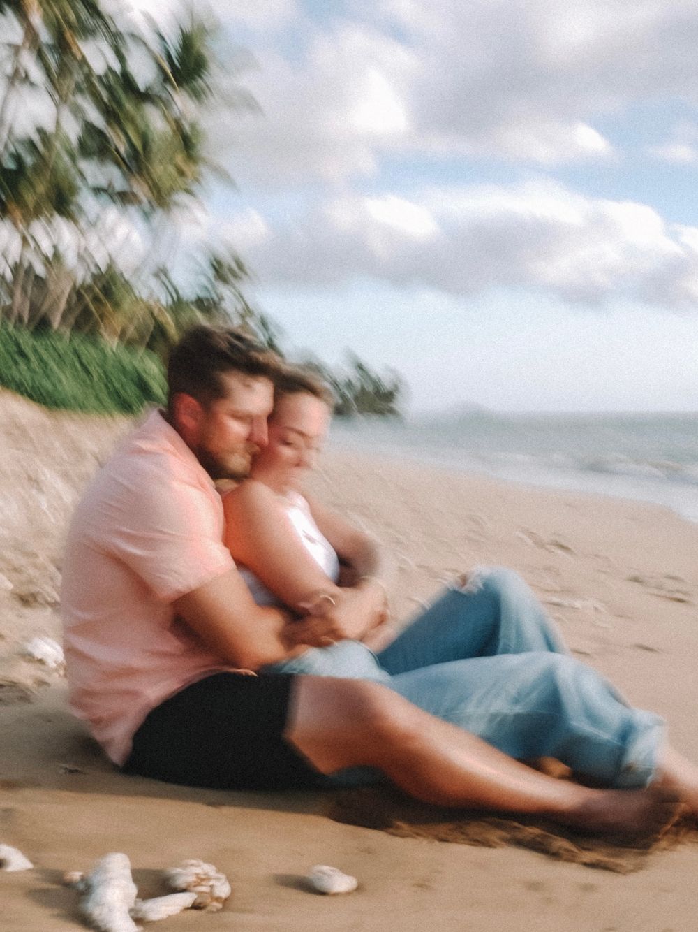 A couple shares playful moments while sitting on a tropical beach with palm trees.
