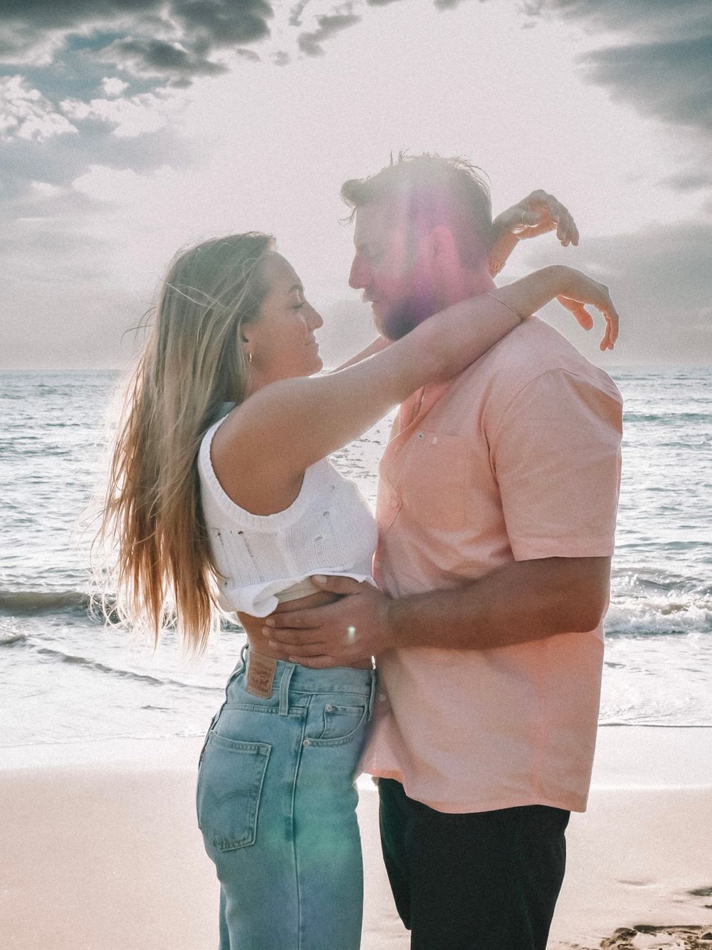 A couple in casual beach attire shares affectionate moments against an ocean backdrop.