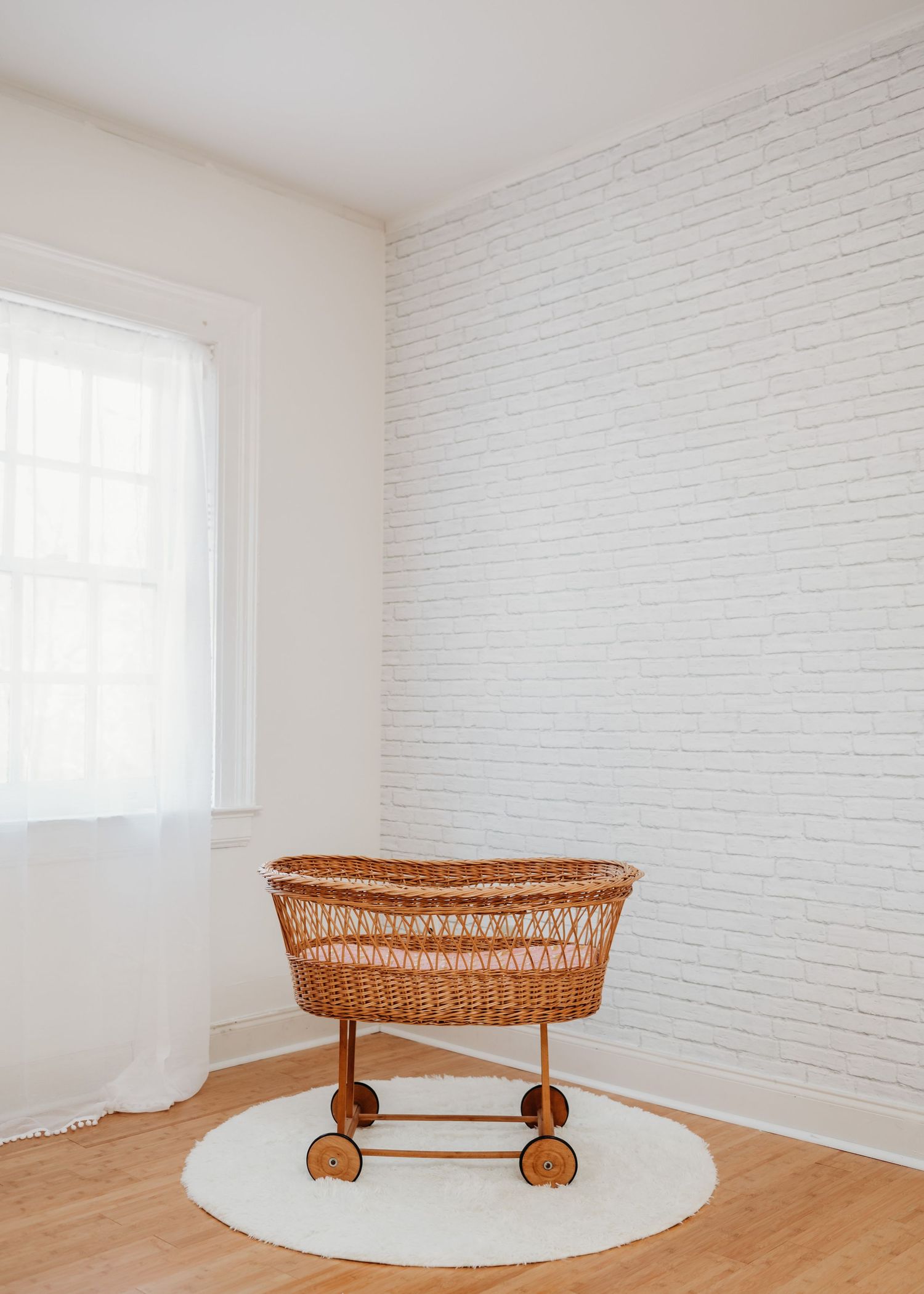 Vintage wicker bassinet on wheels sits on a white round rug against white brick wall in minimalist nursery in Glen Ridge photo studio.