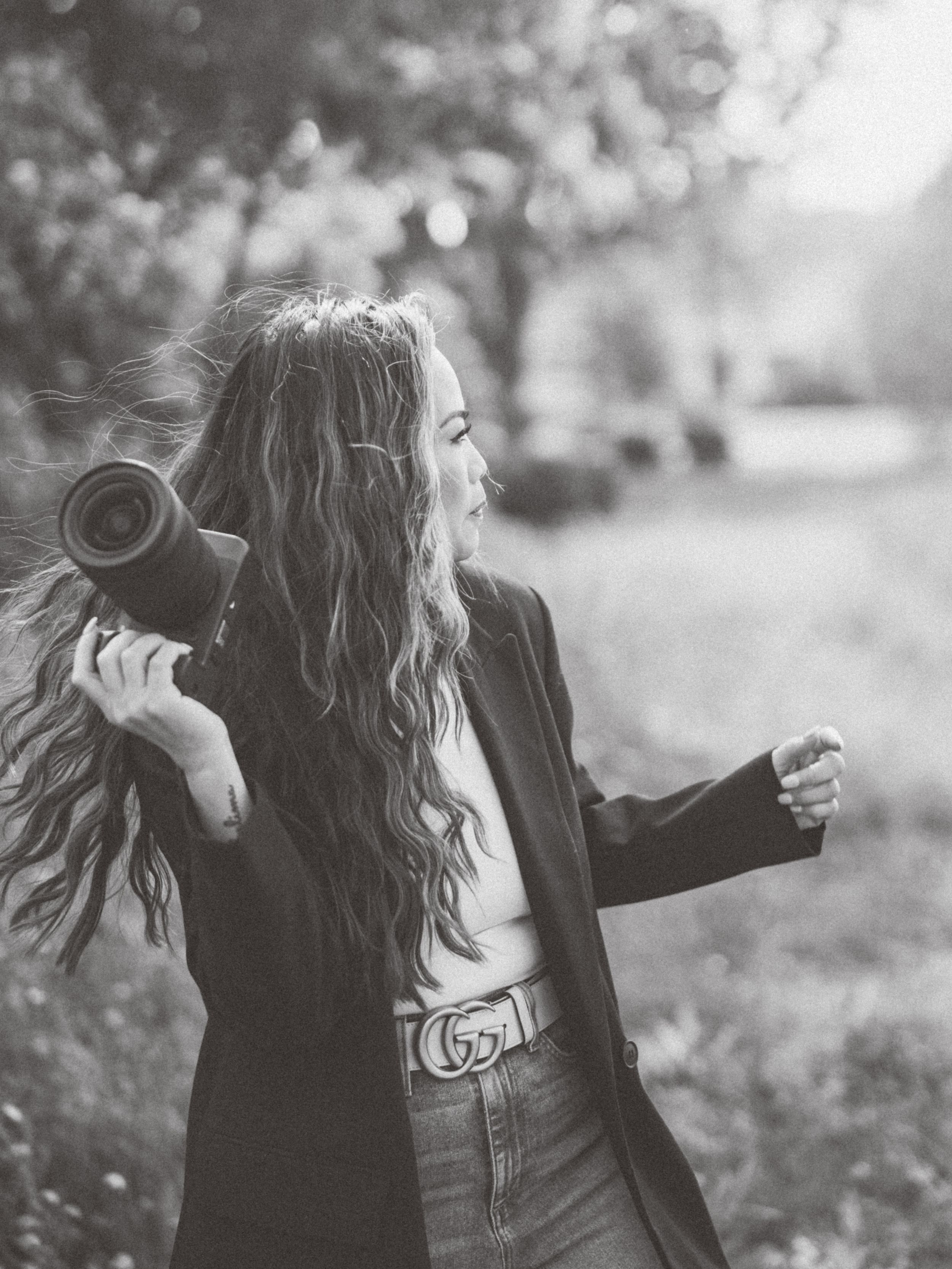 Black and white portrait of photographer with long hair holding camera in natural setting.