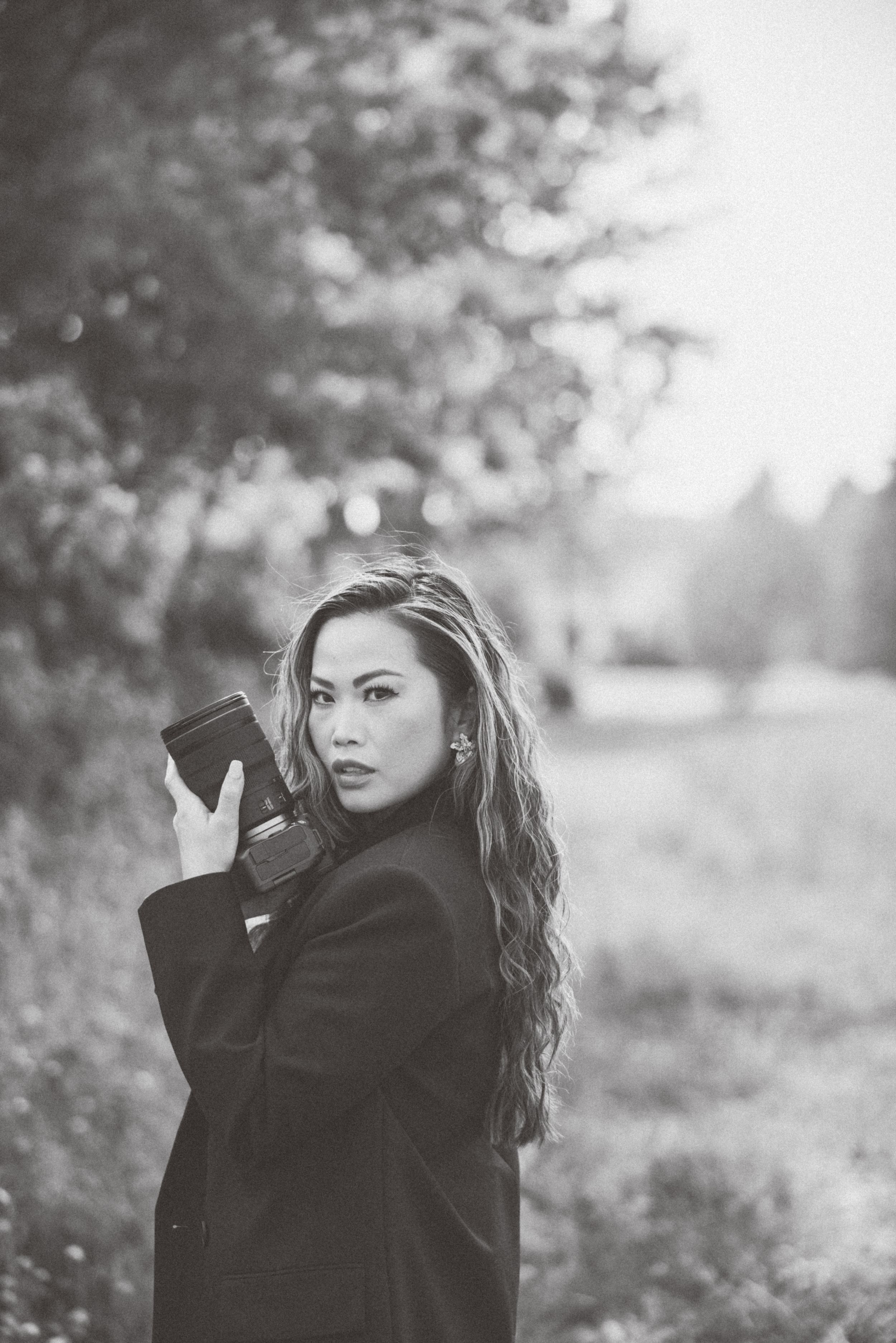 Moody black and white portrait of photographer in black coat against natural backdrop.