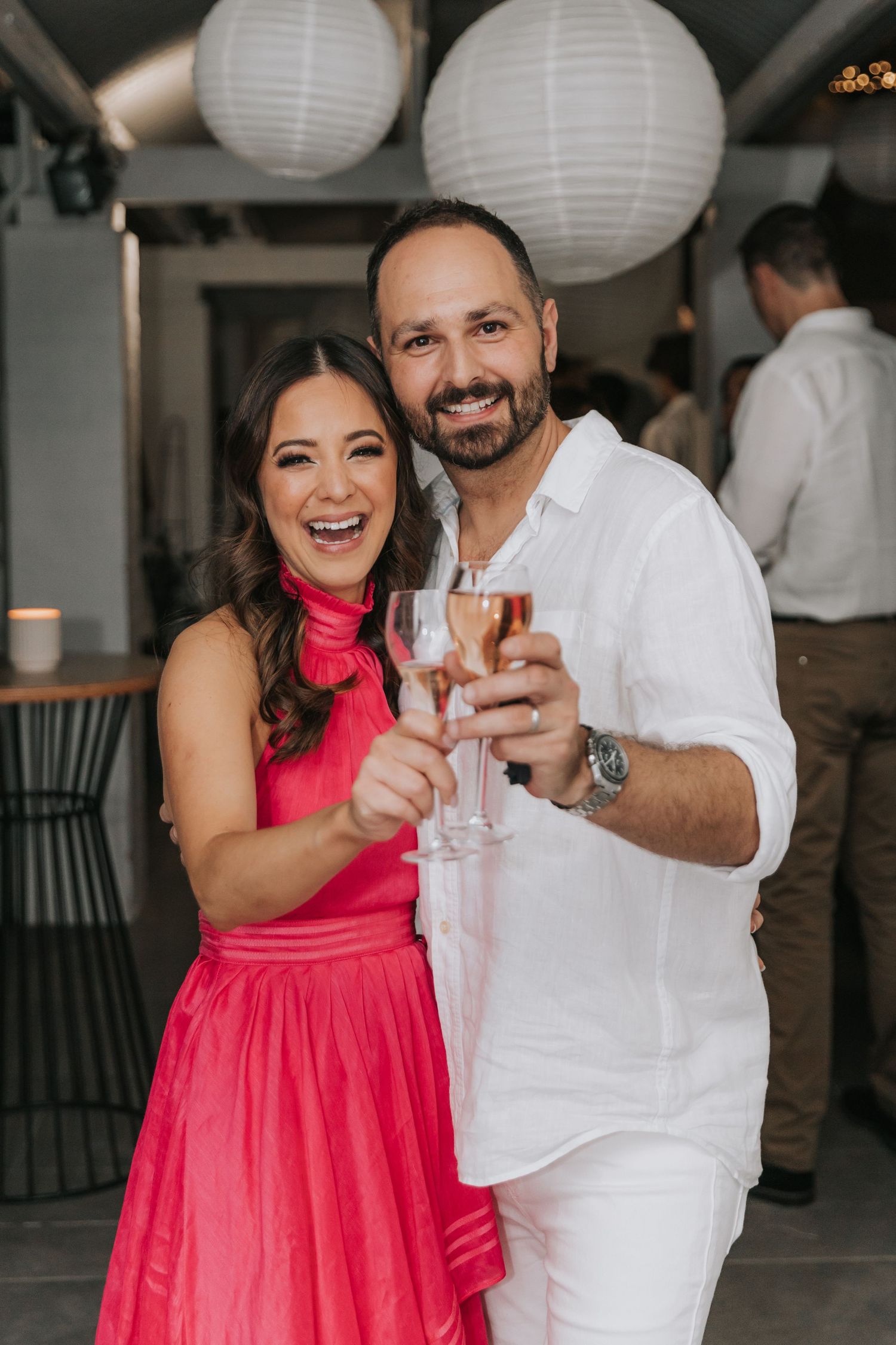 Couple in formal attire sharing drinks at party with white paper lanterns in background.
