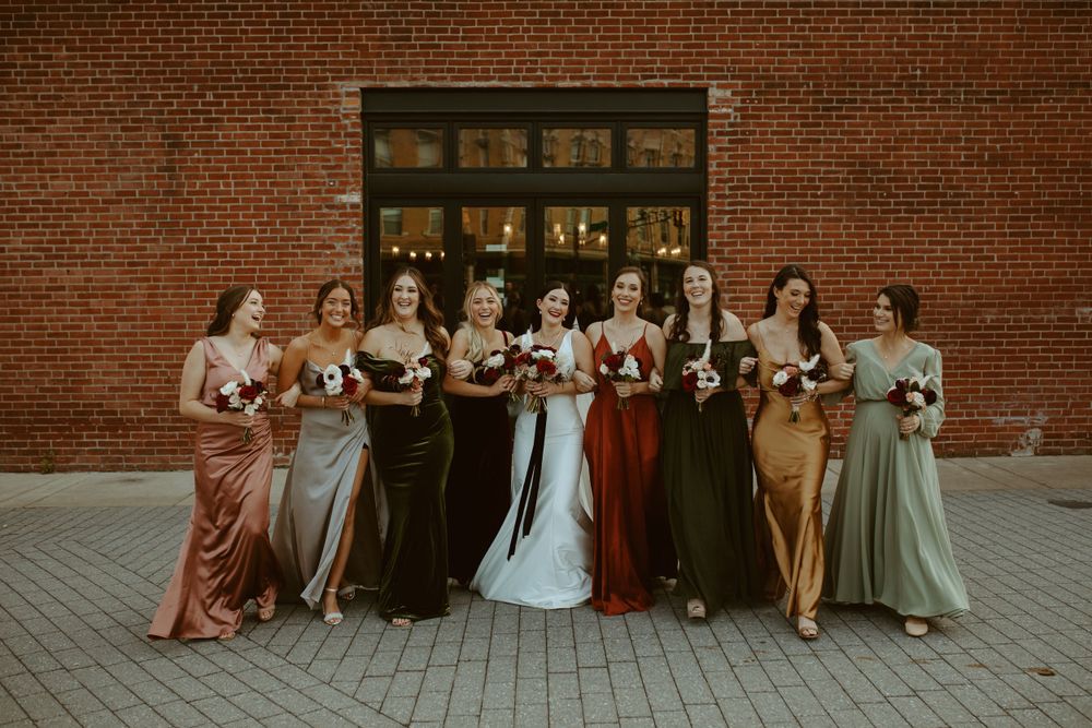 Bride surrounded by bridesmaids in mismatched earth-tone satin gowns outside a brick building.