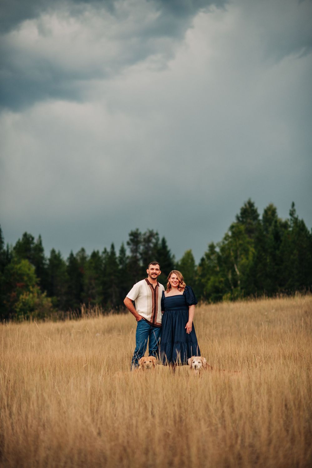 couple standing in a field, dogs are sitting and you can see their heads popping out over the grass, storm looming