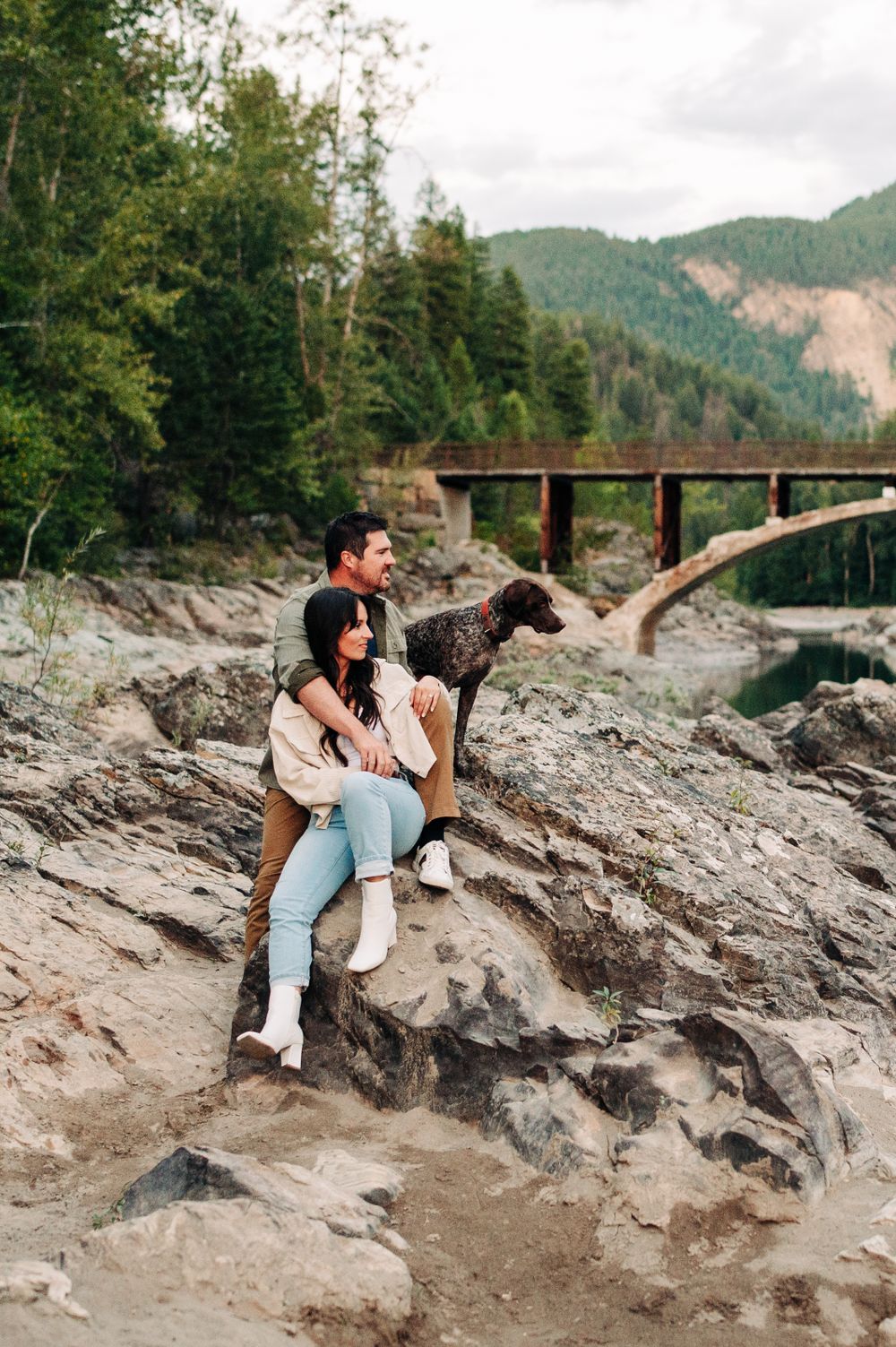 couple sitting on a rocky shore, looking out at the trees with a dog sitting next to them