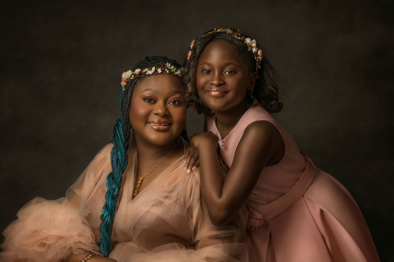 Elegant studio portrait of a mother and her daughter, featuring floral crowns and soft pink dresses against dark backdrop.