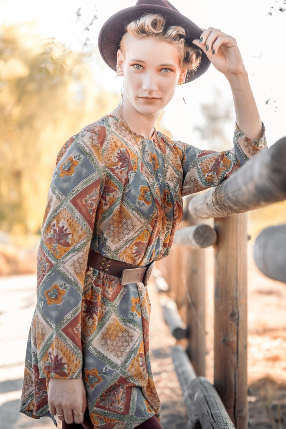 Fashion editorial of a woman in brown tones and a hat, posed on a trail with a wooden fence.