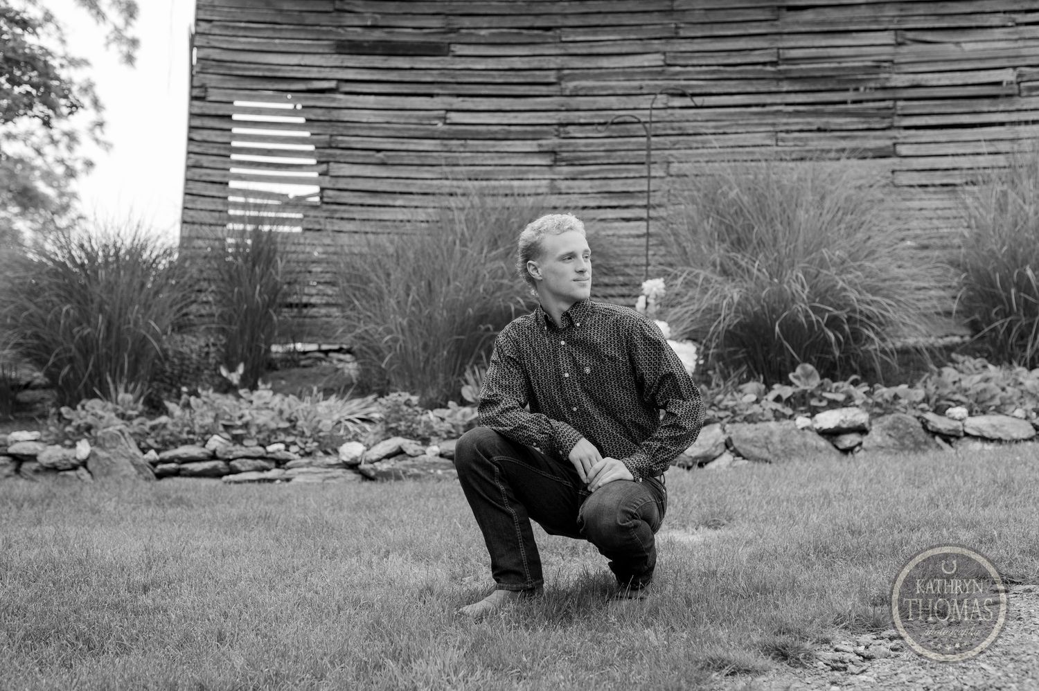 Black and white portrait with weathered wood barn wall and tall grasses in background.