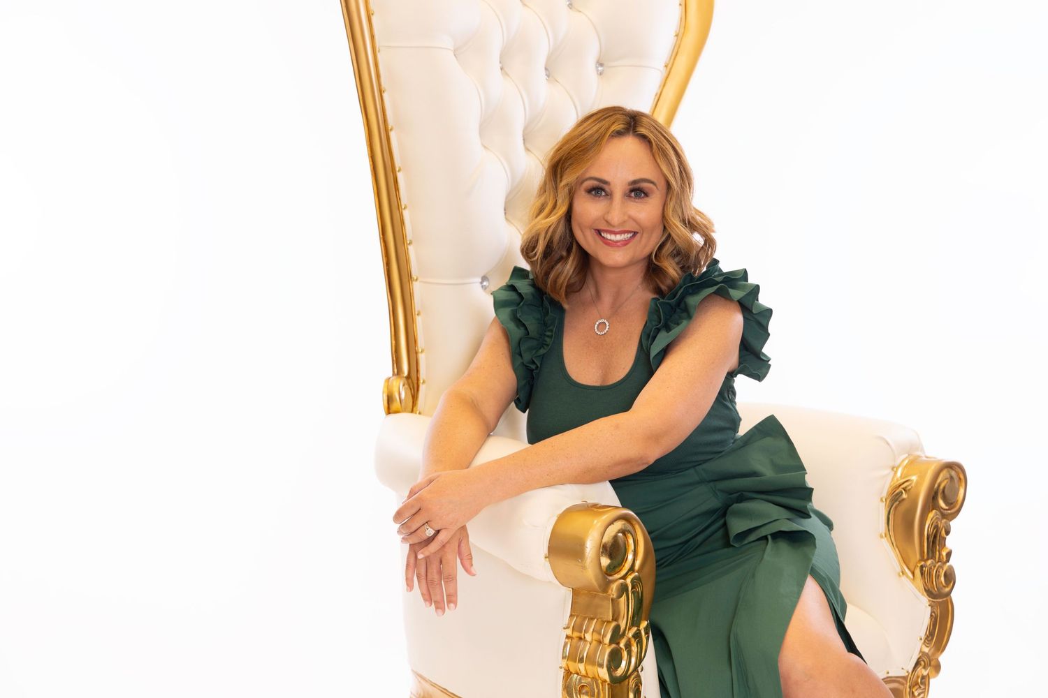Professional portrait of someone in an elegant emerald green dress seated on a white and gold accent chair.