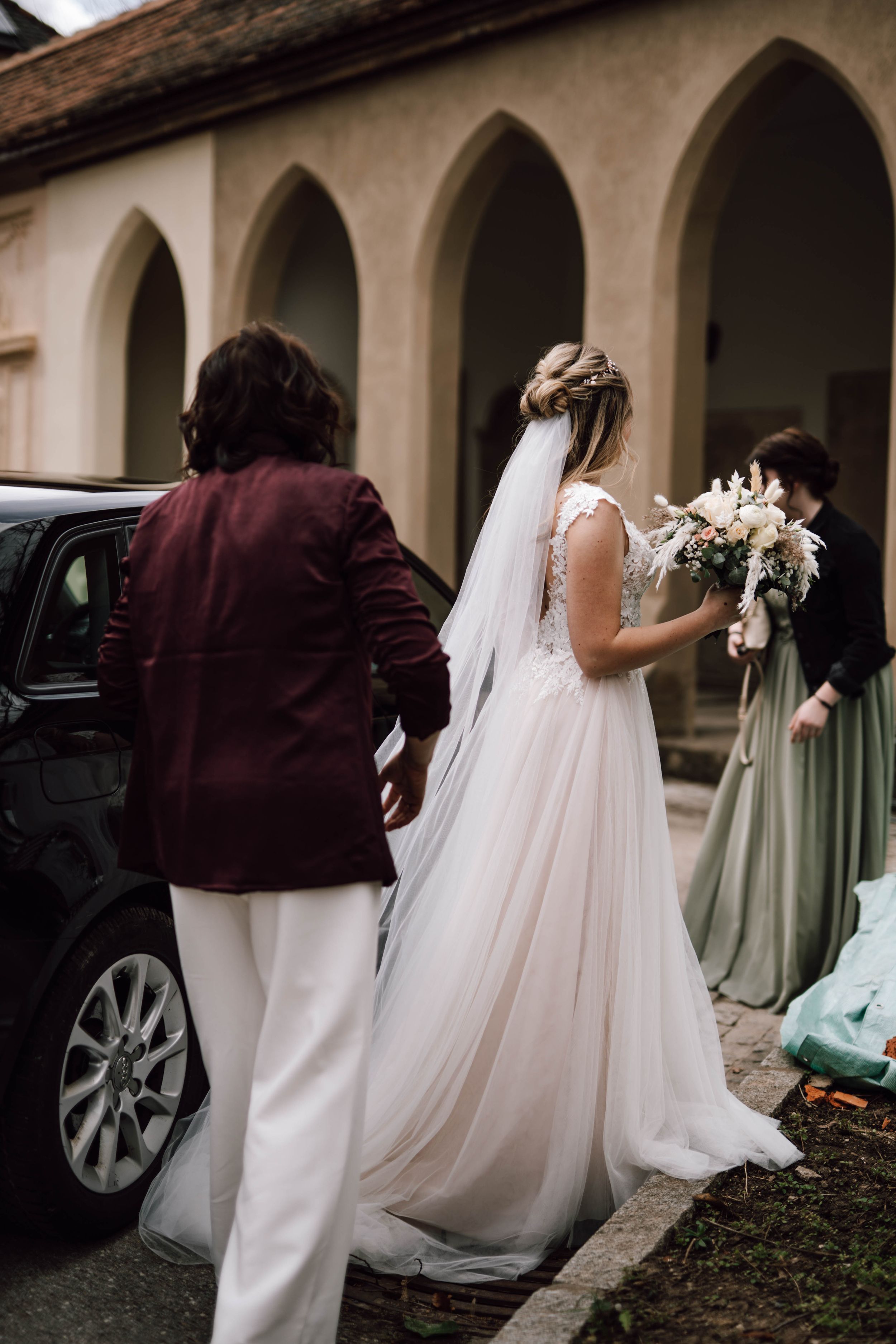 Wedding party walking through arched colonnade with flowing white dress and burgundy accents.